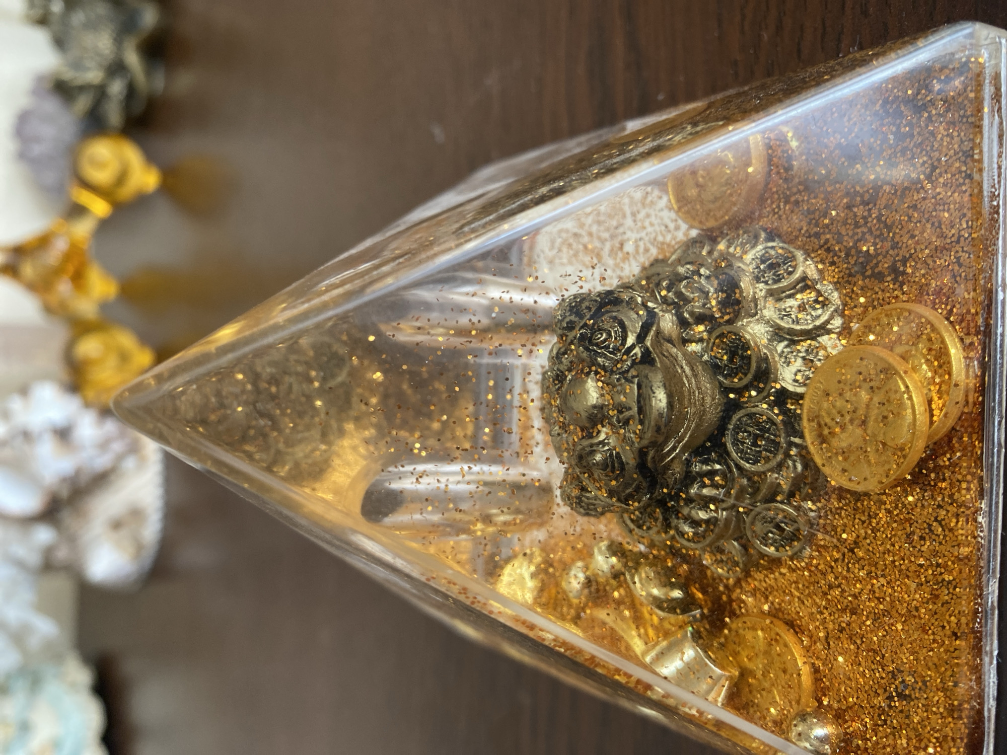 A golden toad figurine sits within a clear pyramid filled with gold glitter and coins. The pyramid is placed on a dark wooden surface, with blurry objects in the background that appear to be decorative items. The scene is indoors, and the lighting suggests it is daytime. The location in Brest, Belarus, is not discernible from the image.