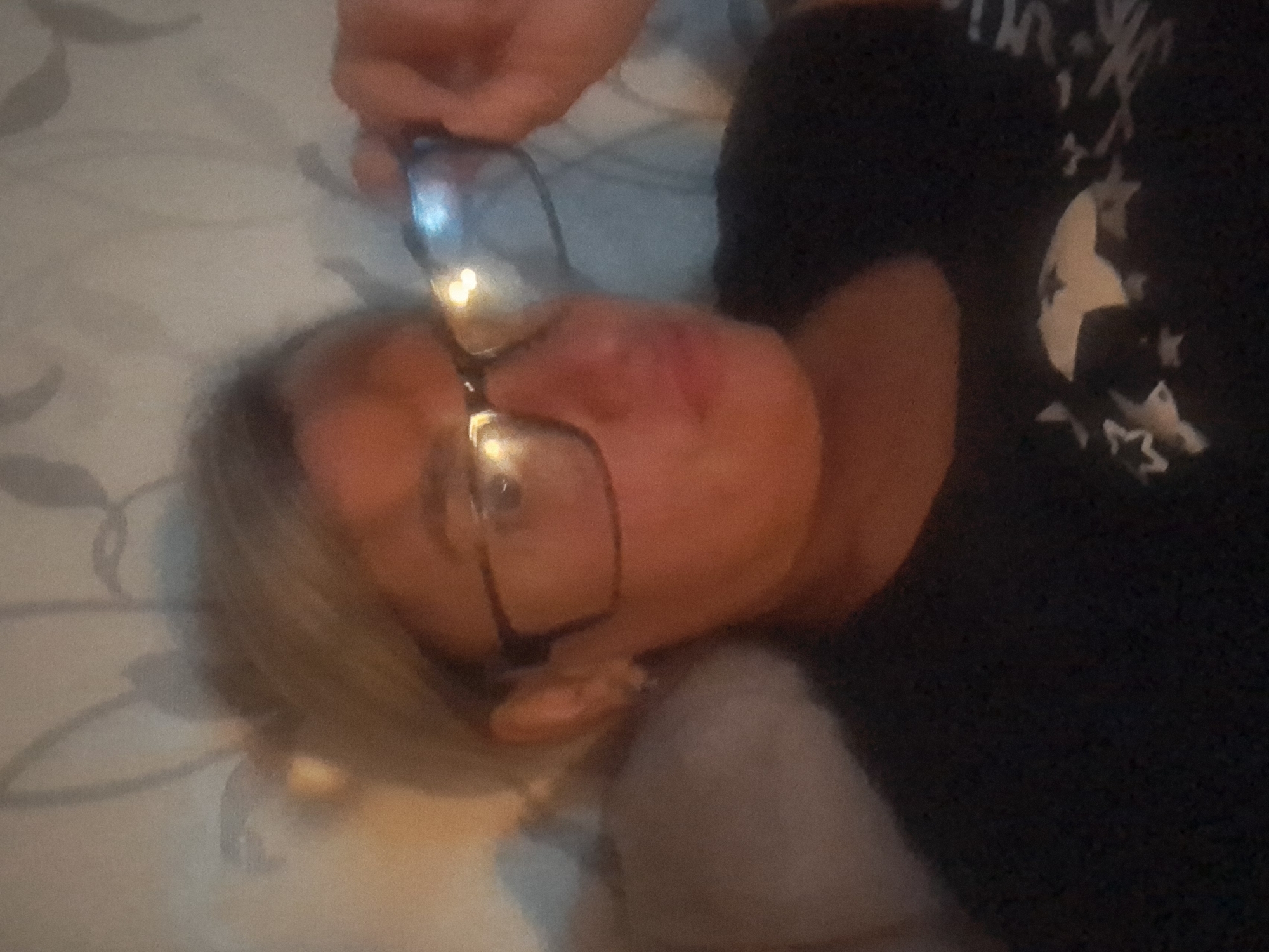 A person, likely an adult female with blonde or light-colored hair, is depicted lying down, possibly on a bed or sofa, in a dimly lit indoor setting. She is wearing dark-rimmed glasses, which exhibit bright glare reflections from an unseen light source. Her face is turned slightly upwards, with her eyes appearing open. A hand is visible near the top right frame of the glasses, though the exact action (e.g., adjusting, holding) is not entirely clear.
She is dressed in a dark-colored shirt, possibly black, adorned with a light-colored, somewhat faded pattern. Visible elements of this pattern include stylized letters or shapes, such as a "W" or "M" on the upper left, and several stars of varying sizes on the right side of the shirt. The background consists of a light-colored, subtly patterned fabric, consistent with bedding or a pillow.
The overall impression is one of relaxation or rest. The dim lighting suggests it might be evening or nighttime. No specific visual cues within the image indicate the provided location context of Elektrostal, Russia. The visible text on the shirt is too indistinct to be fully legible beyond generalized shapes and stars.