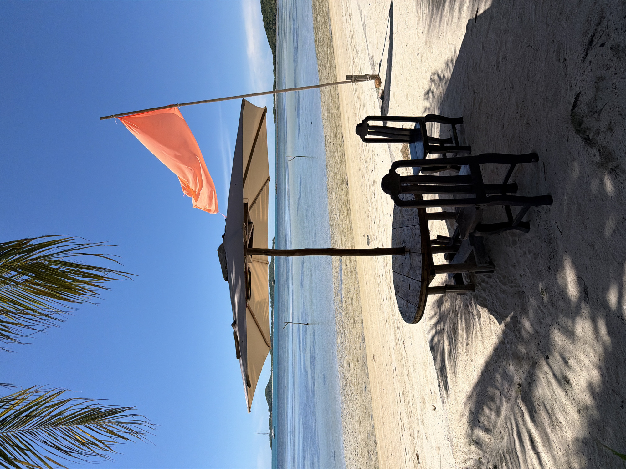 The image depicts a tranquil beach scene in Baan Thong Krut, Thailand. In the foreground, a round wooden table with two dark wooden chairs sits beneath a large, light-colored umbrella, casting shadows on the sand. The umbrella is positioned to provide shade over the seating area. To the right of the umbrella, a bamboo pole stands vertically, supporting a bright orange flag that flutters in the breeze.
The sandy beach gently slopes down to the calm, clear blue water. In the distance, the sea is dotted with a few small boats, and a lush green island with a distinctive peak is visible on the horizon. The sky is a brilliant, cloudless blue, characteristic of a sunny day. Palm fronds hang from the top left of the frame, adding to the tropical ambiance. The overall atmosphere is one of peaceful seclusion and natural beauty.