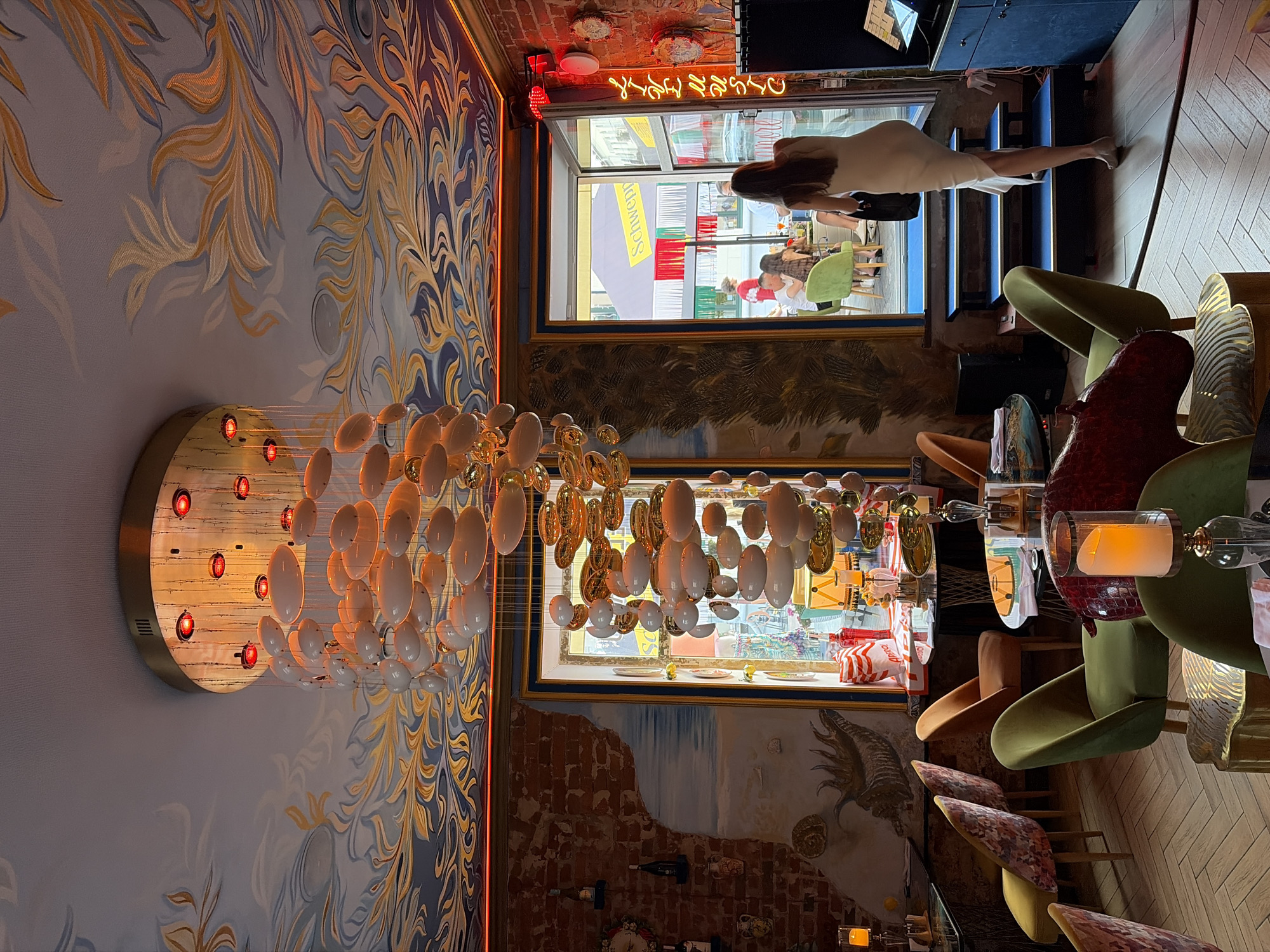 The image captures the interior of a restaurant or bar in Minsk, Belarus. The scene is richly decorated with a vibrant, patterned ceiling featuring swirling gold and blue motifs. A large, multi-tiered chandelier with numerous oval-shaped, translucent light fixtures hangs prominently in the center. The walls are a mix of exposed red brick and painted murals, one depicting a seascape and another with abstract designs.
Several tables and chairs are arranged throughout the space. The chairs have varied upholstery, including floral patterns and solid green velvet. One table in the foreground holds a lit candle in a glass hurricane lamp.
A woman in a cream-colored dress is walking away from the camera towards an open doorway. Through the doorway, other patrons are visible sitting at tables outdoors, with a Schweppes umbrella providing shade. A neon sign spelling "LIVE MUSIC" is mounted vertically on the brick wall near the entrance. The floor is laid with light-colored wooden planks in a herringbone pattern. The overall ambiance suggests a lively and artistic establishment.