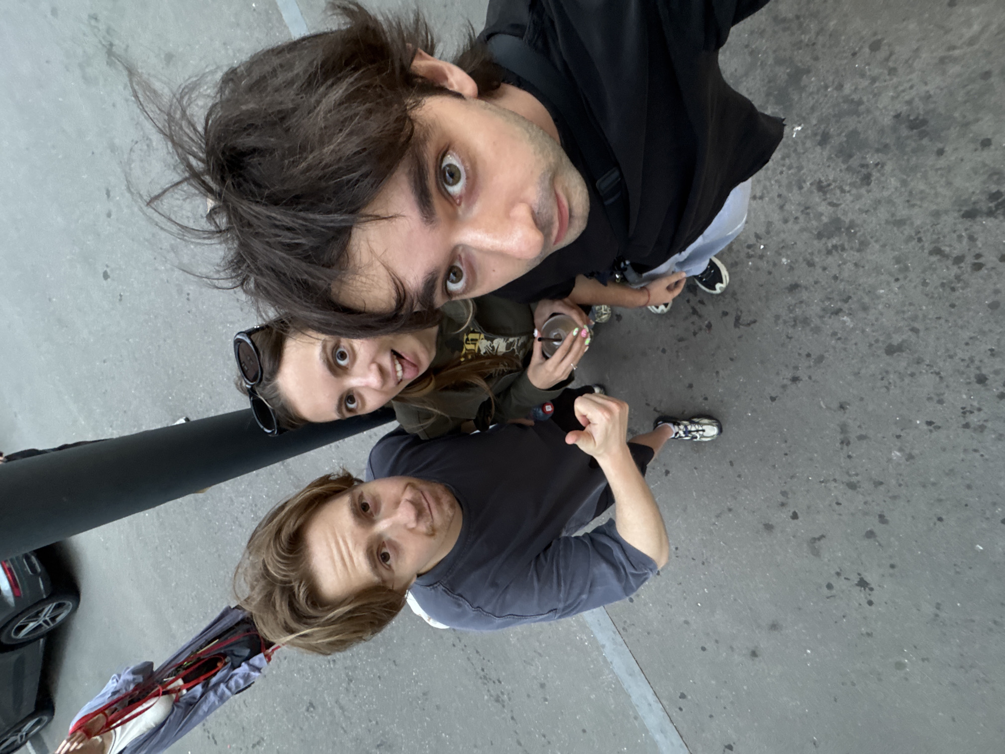 The image shows three people, two men and one woman, taking a selfie from a high angle. The first man, in the foreground, has light brown hair and is wearing a dark grey t-shirt. He is looking directly at the camera with a slight smile and his right arm is raised with his fist clenched. To his left and slightly behind him is the woman, who has dark brown hair and is wearing sunglasses pushed up on her head. She is looking at the camera with wide eyes and a surprised expression, holding a drink with a straw. Behind her and to the left is the second man, who has dark brown hair and is wearing a black t-shirt. He is also looking at the camera, with a surprised expression. The background appears to be a paved area, likely a street or sidewalk, with some dark splatters on the ground. A dark pole or railing is visible on the left side of the image, and part of a car is visible in the lower left corner. The lighting suggests it is daytime. The overall mood is casual and playful. No visible text is present. The location context provided is Moscow, Russia.