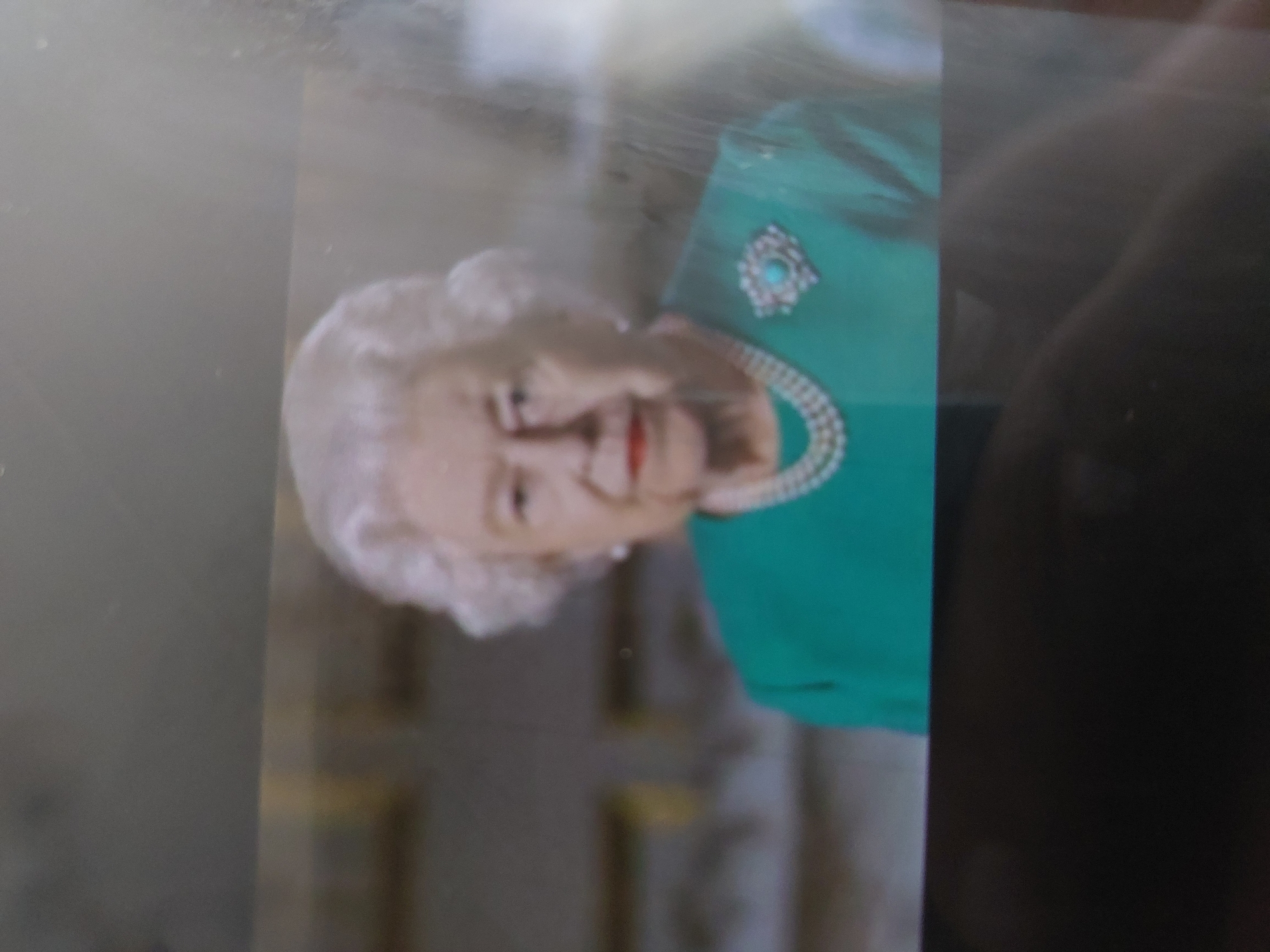The image shows a photograph of Queen Elizabeth II, wearing a green dress with a bejeweled brooch and a pearl necklace. It's not possible to identify the location of the image or the city of Tsarevo, Bulgaria, within the photograph itself. The photograph appears to be of a print or screen, as evidenced by the glare and surface textures.