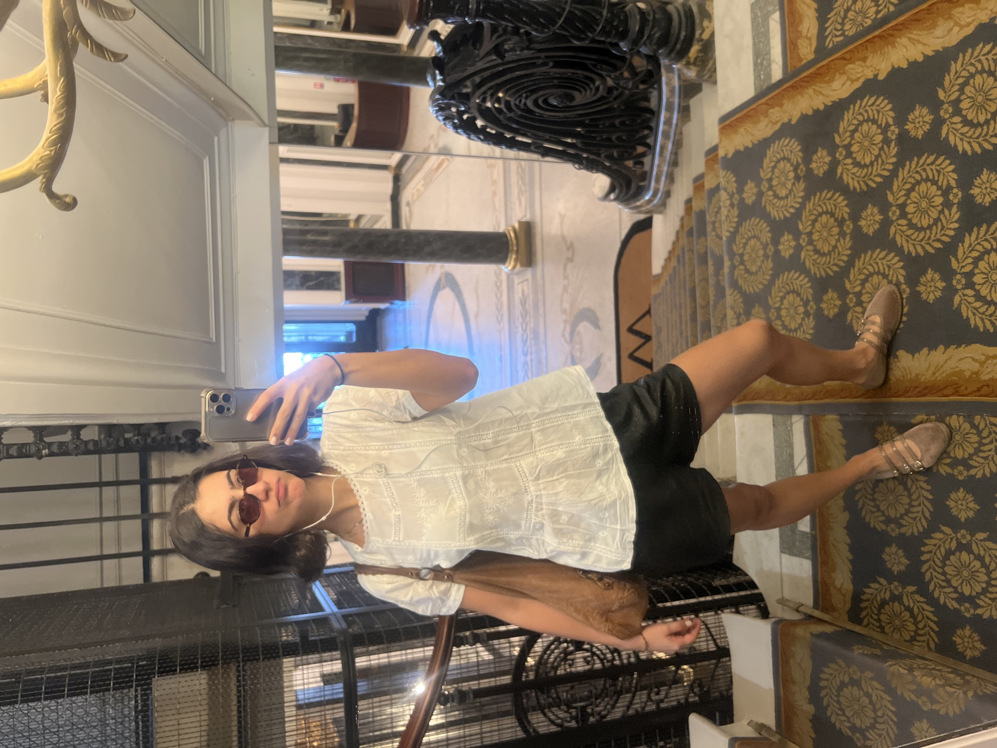 The image shows a woman taking a selfie in what appears to be an ornate building in Paris, France. She is standing on a carpeted staircase with a decorative floral pattern. The architecture features marble floors, a wrought-iron staircase railing, and columns. The reflection in the mirror behind her shows a chandelier and additional architectural details. The woman is wearing sunglasses, a white top, and black shorts. She is holding a smartphone to take the photo. She has a brown purse over her shoulder.