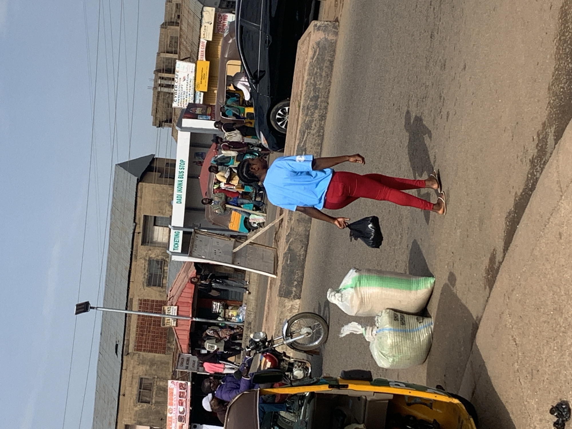 The image depicts a street scene in Gura Top, Nigeria, likely a bus stop area. A young woman in a blue t-shirt and red leggings walks across the asphalt road carrying a black plastic bag. To her left, two large sacks are placed on the ground, one green and white, and the other off-white. A yellow and green motorized tricycle, commonly known as a "Keke Napep," is partially visible on the left edge of the frame. In the background, various commercial establishments line the street, including one with a "TICKETING" sign and another labeled "DADI IKOWA BUS STOP." Several people are seen milling about, some near a motorcycle and others near the storefronts. A black car is parked further down the road. The environment appears to be a bustling urban area with a clear sky.