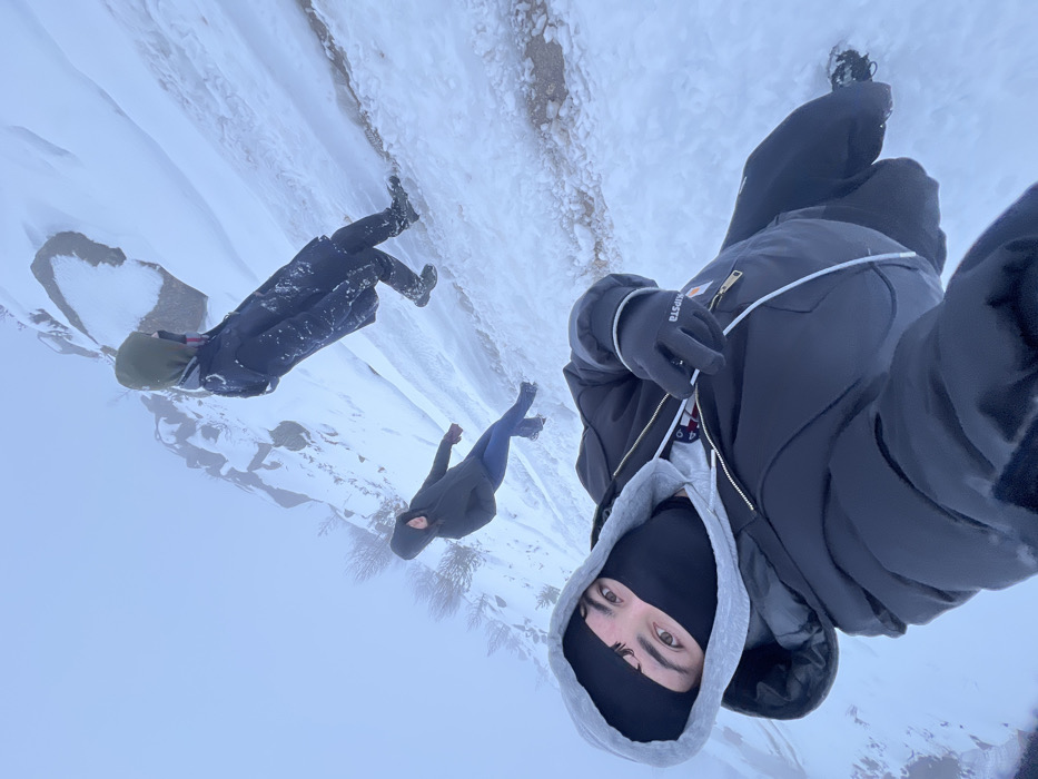 The media file captures a snowy outdoor scene featuring three individuals on a hillside or mountain slope. The perspective is a wide-angle, selfie-style shot, looking out over the landscape.
In the foreground, positioned towards the lower right, is the person taking the photograph. They are wearing a black balaclava, revealing only their eyes, which look directly towards the camera. Their attire consists of a grey hooded sweatshirt underneath a dark, likely black or charcoal, puffy winter jacket with gold zippers and a distinctive white cord detail. A black glove, clearly showing the brand "KIPSTA" in white text, is visible on their left hand.
Further into the scene, to the left of the foreground subject, stands a second person. They are dressed in a dark jacket, possibly with a green hood, dark trousers, and boots, standing on the snow-covered ground. Behind and slightly to the left of them, a third individual is also visible, clad in similar dark winter clothing.
The setting is dominated by a thick layer of snow covering a sloping terrain, suggesting a mountain path or hillside. The background is a bright, hazy expanse, indicating either an overcast sky or fog, which blends with the snow in the distance. Sparse, dark vegetation or bushes can be faintly seen through the mist further down the slope. The scene conveys a cold, wintry environment, consistent with hiking or exploring snowy landscapes in a region like Vila Real, Portugal.