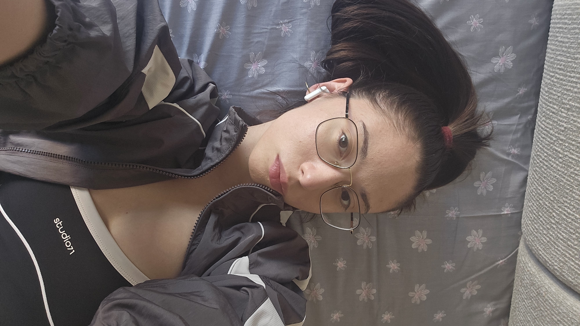 A young woman lies on a bed, wearing a black and white sports bra under an unzipped gray and white tracksuit jacket. She has dark hair pulled back in a ponytail with a red hair tie. She is wearing eyeglasses and has a white earbud in her left ear. The bed has a gray sheet with a floral pattern. Part of a gray couch is visible on the right side of the frame. The image does not provide location information; thus, I cannot determine if the location is Stara Zagora, Bulgaria.