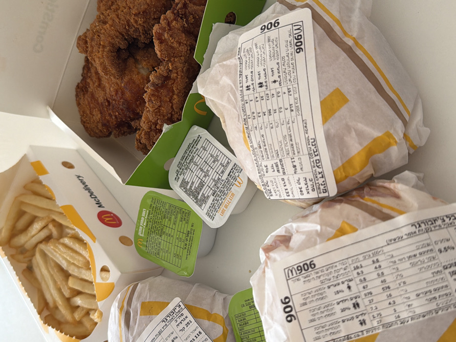 The image presents a top-down view of a McDonald's fast food meal, consistent with a delivery order, laid out on a clean, white surface. This meal originates from a McDonald's in Tel-Aviv, Israel, identifiable by the "McDelivery" branding and Hebrew text on all packaging.
The items visible include:
* An open white cardboard box containing french fries, prominently displaying the McDonald's logo and "McDelivery" in red and black text.
* An open cardboard box, lined with green, holding several pieces of breaded fried chicken.
* Three food items wrapped in McDonald's branded white and yellow striped paper. Two of these are prominent, each sealed with a white adhesive label featuring detailed nutritional information in Hebrew and the code "906" or "M906." These wrapped items are likely burgers or sandwiches. A third, partially obscured wrapped item is visible on the left.
* Two small plastic sauce cups. One features a green lid, indicating a specific sauce (likely sweet and sour, with Hebrew labeling and nutritional facts). The other has a white lid. Both cups display the McDonald's logo and Hebrew nutritional information.
All packaging is intact and clean. No individuals or external environmental details beyond the white surface are visible.