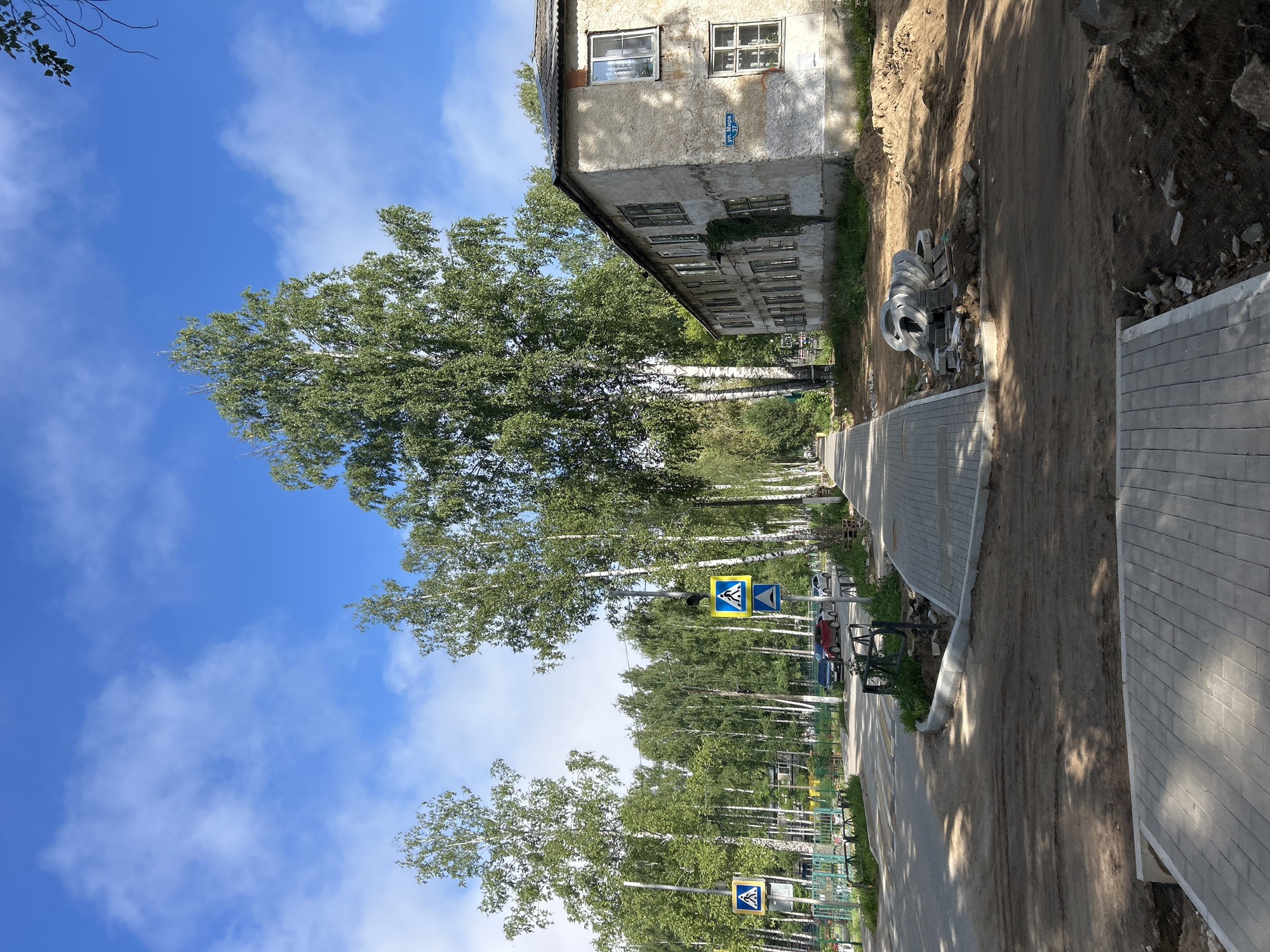 The image depicts a street scene in the city of Chaikovsky, Russia. The foreground shows a newly paved sidewalk made of gray interlocking pavers. To the right of the sidewalk, there is an area of disturbed earth with piles of sand and construction debris, including stacked circular concrete elements. In the middle ground, the paved sidewalk continues along a street lined with tall birch trees. A crosswalk is visible on the street. Several cars are parked or driving on the street. On the right side of the street, an old, two-story apartment building with a weathered facade and numerous windows stands. A street sign with Cyrillic lettering ("ул. Мира") is affixed to the building. The sky is blue with scattered white clouds. The overall impression is of an ongoing urban improvement project in a residential area.