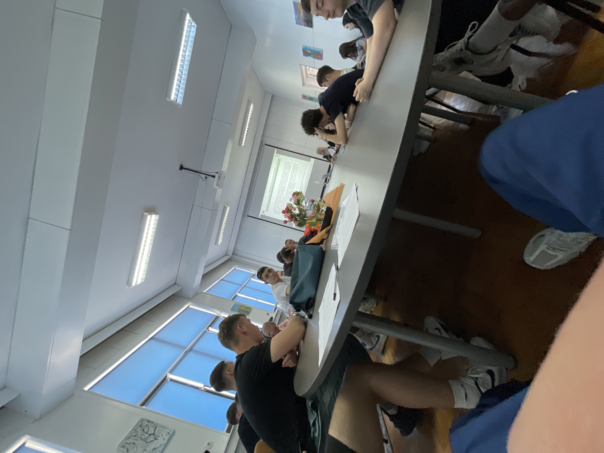 A long table is surrounded by a group of young people in a brightly lit room, likely a classroom. The location could be Zlatograd, Bulgaria. The young people are sitting and appear to be engaged in a lecture or presentation, with a projection screen visible at the end of the table displaying text. There is a vase of flowers on the table near the screen. The people are dressed casually and appear to be focused on the presentation.