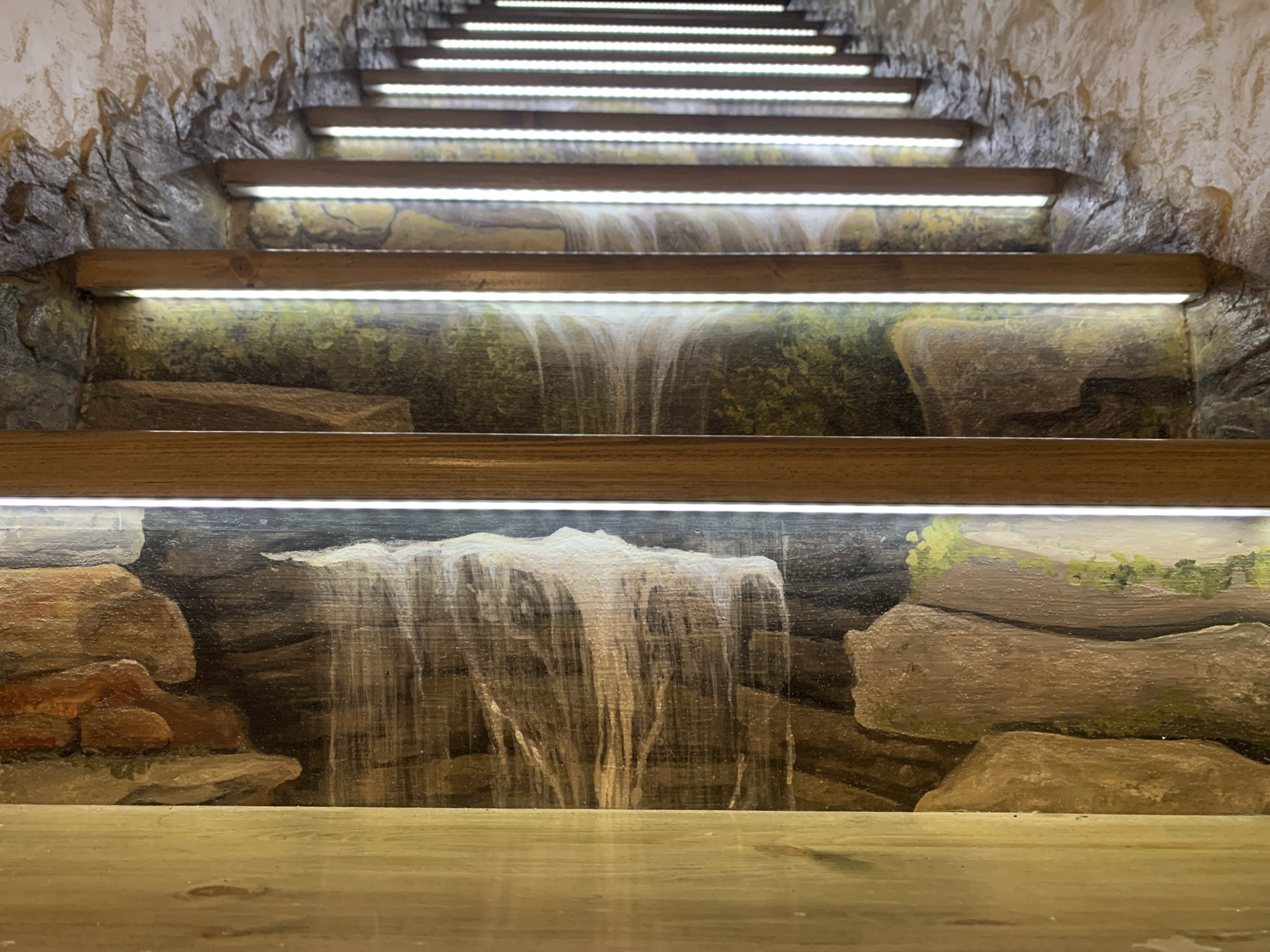 A close-up shot of a set of interior steps painted with a waterfall scene, presumably located in Severodvinsk, Russia. The steps are illuminated by what appears to be LED light strips positioned underneath each tread. The risers of the steps are painted to depict a continuous waterfall and rock formation scene, creating a faux-outdoor environment. The steps themselves are made of wood and have a clear finish. The surrounding walls are textured to resemble stone or a cave.