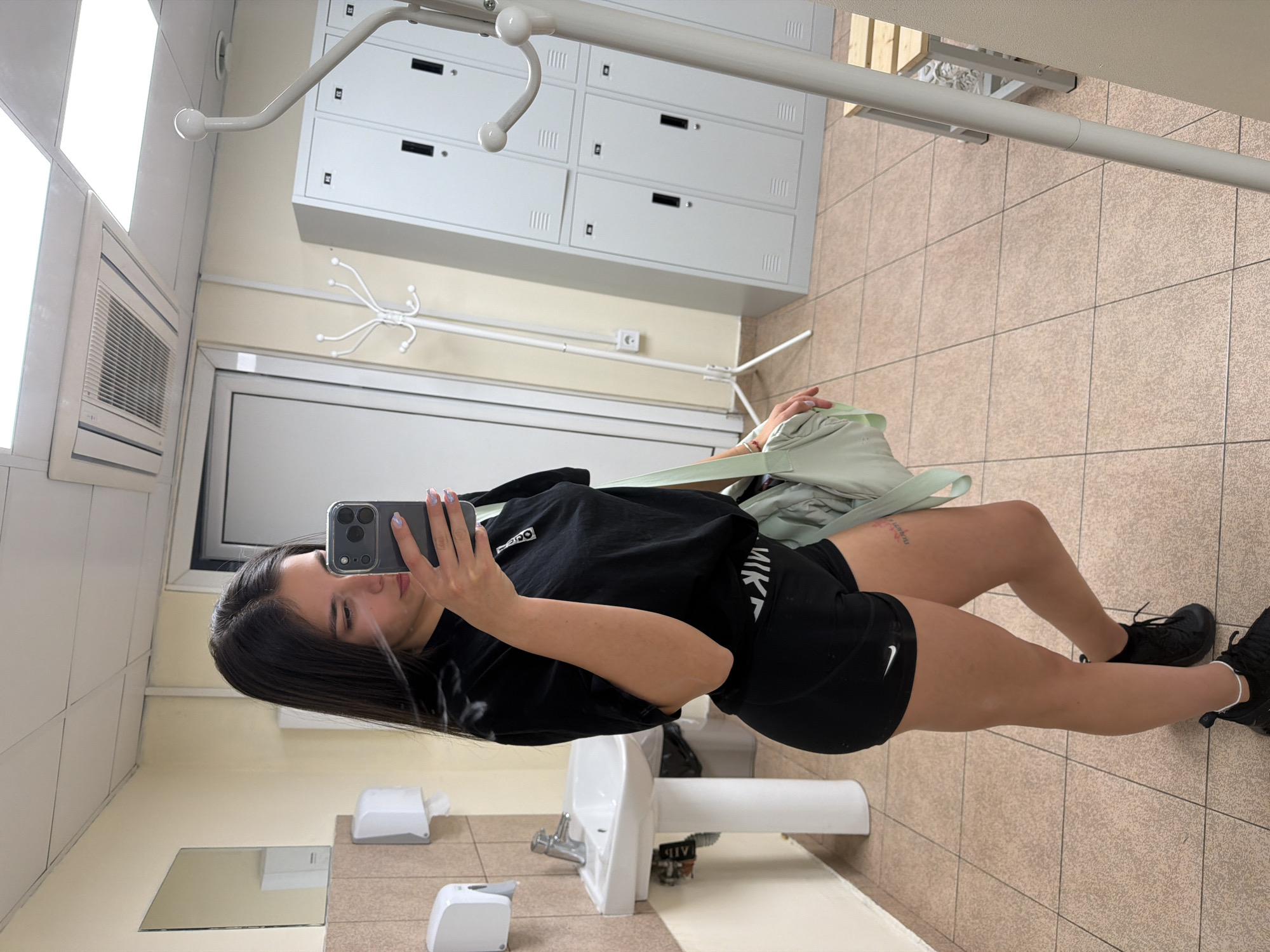 A young woman stands in what appears to be a locker room or public restroom, as suggested by the presence of lockers, a sink, and hand-drying equipment. The woman is taking a selfie with her phone, which she holds up in her left hand. She is wearing a black t-shirt and black shorts with a white Nike logo, along with black athletic shoes. The background includes beige-tiled walls and floors, white lockers, and hand-drying equipment. It is not possible to ascertain if this image was taken in Gabrovo, Bulgaria.