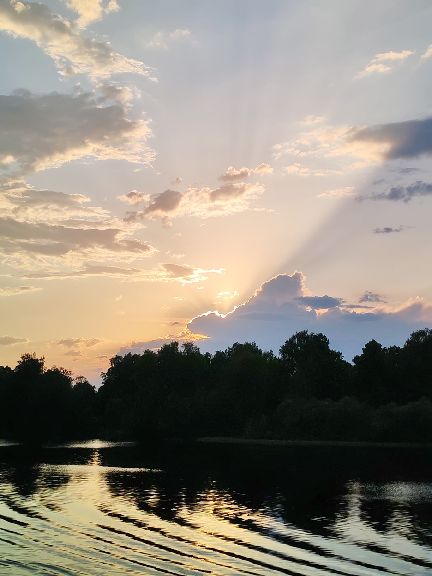 The image captures a sunset over a body of water, likely a river or lake, near Krasnaya Gorka, Russia. The sky is filled with clouds illuminated by the setting sun, creating a mix of golden, orange, and light blue hues. Sunbeams are visible, radiating from behind a large cloud. In the middle ground, a dark treeline silhouettes against the bright sky. The water in the foreground reflects the colors of the sky and the sun's light, creating patterns of light and dark ripples. The water's surface shows distinct wave patterns suggesting some form of movement. No people or distinct objects are present.
