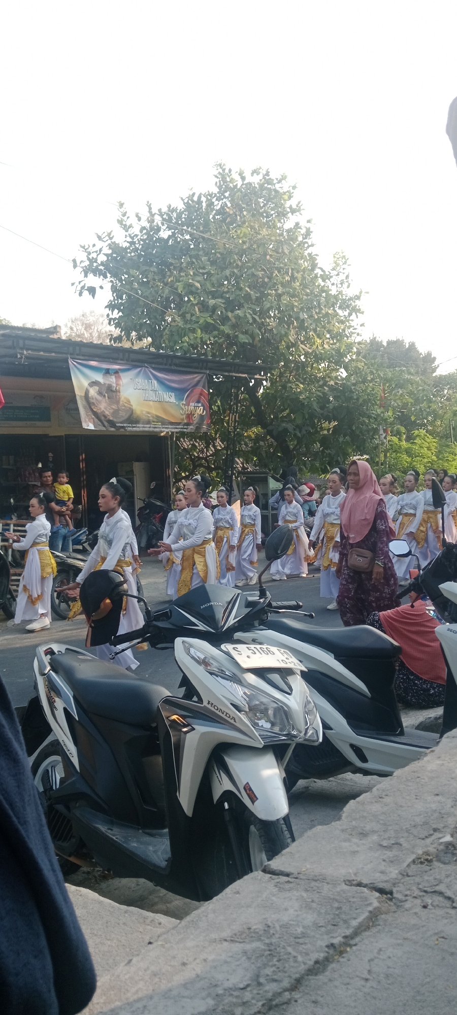 The image captures a street scene in Soko, Indonesia, likely during a procession or cultural event. In the foreground, two parked motorcycles are visible. The left motorcycle is predominantly black, while the right one is white with black accents. The white motorcycle has a license plate that reads "S 5166 FO".
Behind the motorcycles, a group of women in traditional attire are walking. They are wearing white tops and gold skirts or sashes, with their hair styled up. One woman in the front right is wearing a pink hijab and a patterned dress. Several younger women and girls are also part of the group, dressed in similar white and gold outfits.
In the background, a banner hangs above a building, displaying text and an image, though the details are not entirely clear. Trees with lush green foliage line the street, and the sky is bright, suggesting it is daytime. The overall atmosphere appears to be one of a public gathering or celebration.