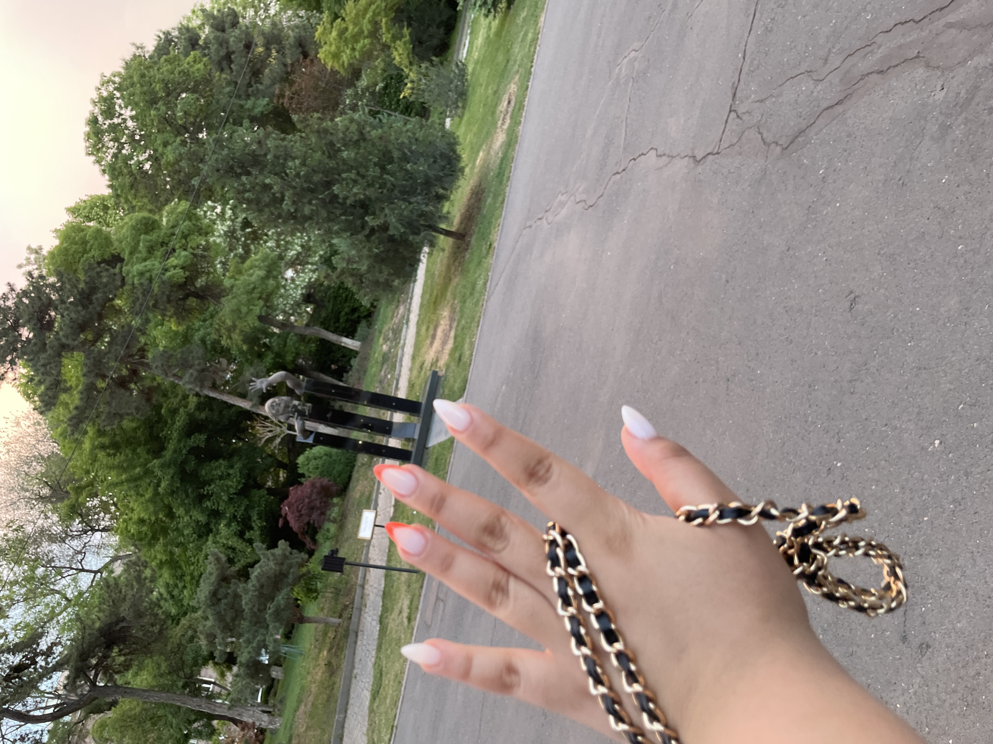The image is taken outdoors in Yerevan, Armenia. The foreground is dominated by a hand with distinctive nail art, wrapped with a black and gold chain. The background features a cracked asphalt path and green foliage, suggesting a park or garden setting. A sculpture composed of three figures stands on the lawn.
