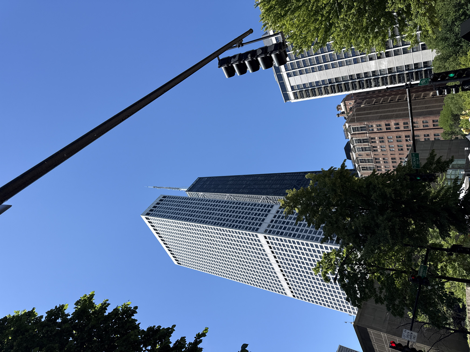 This media file depicts an upward-angle view of a city streetscape with clear blue skies. The primary subjects are several tall buildings with distinct architectural styles, indicative of an urban environment. In the foreground, a black traffic light pole with multiple signal heads stretches diagonally across the frame. Behind it, a predominantly white skyscraper with a grid-like pattern of windows dominates the mid-ground. Further back, a darker skyscraper with a more complex geometric facade is visible, and beyond that, a brick building with numerous windows. Lush green trees are interspersed among the buildings, their foliage creating a contrast with the man-made structures.
The time of day appears to be daytime, given the bright sky and shadows. The weather seems clear and sunny. The presence of traffic lights, street signs, and towering skyscrapers strongly suggests an urban setting, specifically identified as Chicago, USA. No people are visible in the frame. The activity is static, focused on the architectural landscape and urban infrastructure.
Notable details include a street sign visible on a pole that reads "E CHICAGO AVE 800 N," confirming the location. Another street sign is partially visible and appears to read "URBAN ST 300." A traffic signal shows a green light for a bicycle lane, and another shows a red light. The overall impression is one of a bustling city on a clear day, with a focus on the verticality of the architecture.