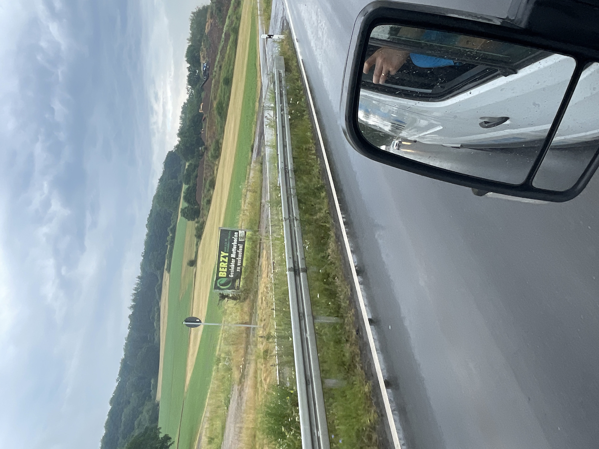 The image is a forward-facing exterior shot of a white van's side mirror, taken from inside the van. The mirror reflects the van's exterior, the road, and the surrounding landscape under an overcast sky. In the distance, there are rolling hills with agricultural fields in shades of green and yellow, and a forested area. A road runs along the edge of the fields, with a metal guardrail. A billboard advertising "BERZY" and "Gesiebter Mutterboden zu verkaufen!" (Sifted topsoil for sale!) is visible on the side of the road. The road surface is wet, indicating rain. In the mirror's reflection, the hand of the van's driver, wearing a ring, is visible, along with a blue shirt. Further down the road, a white car is seen through the van's window. The scene appears to be a rural area outside Wartenberg, Germany.