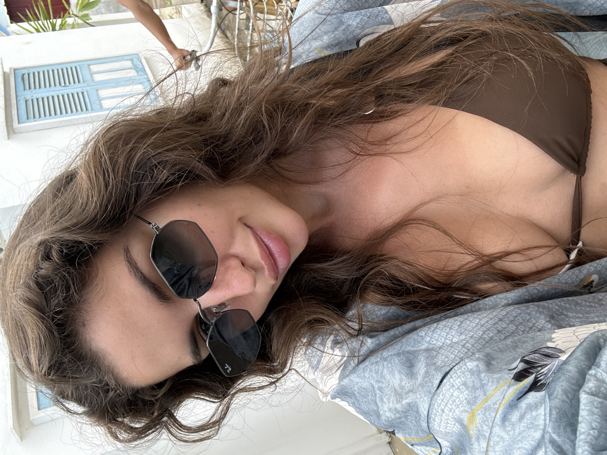 A woman is lying down in what appears to be a bed or cushioned area. She has long brown hair, and is wearing sunglasses and a brown bikini top. The bedding is blue with a crane design. Behind her, partially visible, is a white wall with a small blue-shuttered window. A person's arm is visible reaching towards the window. A bicycle is also partially visible. The city of Unknown, Indonesia is not relevant as location cannot be determined from image.