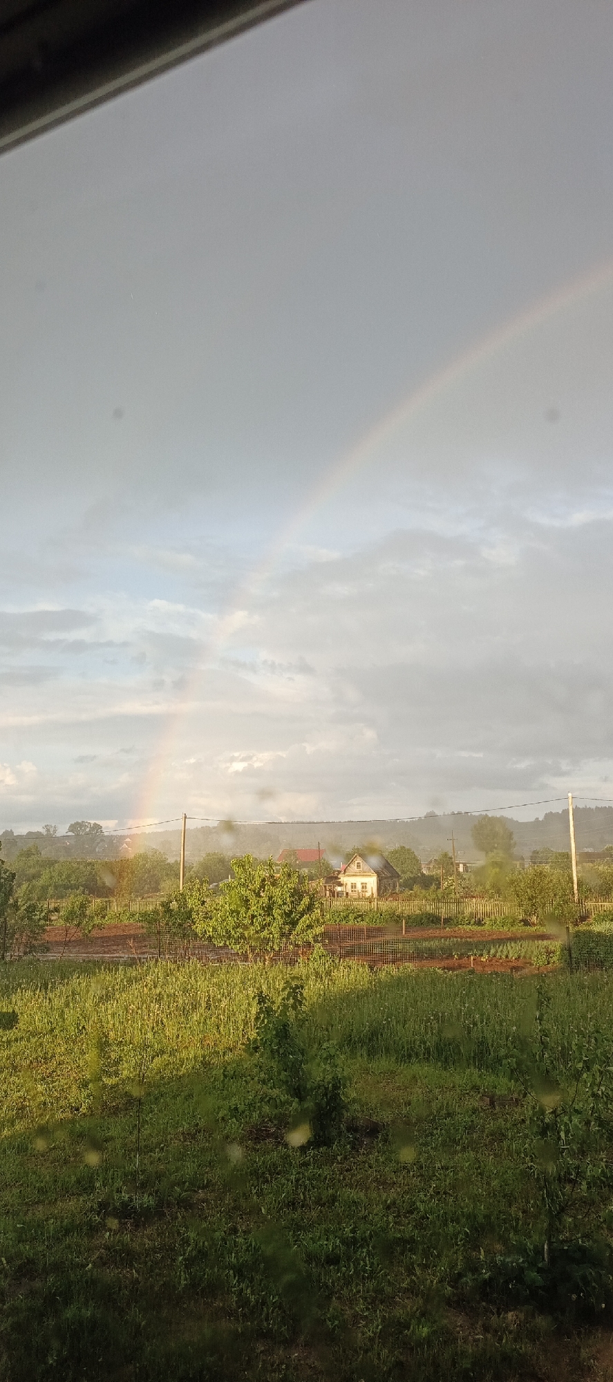 The image depicts a rural landscape in Большой Букор, Russia, featuring a rainbow in the sky. A white house with a red roof is visible amidst greenery and gardens, with wooden fences. A green field occupies the foreground. The photograph appears to be taken through a window, with raindrops visible on the glass. A utility pole is present near the house. Overcast skies are above the village landscape.