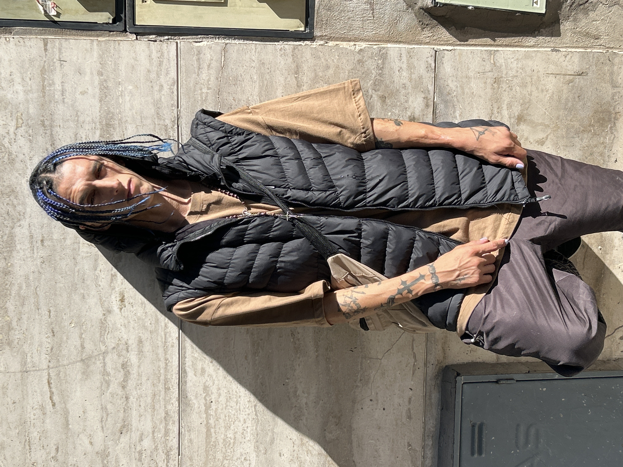 A full-body shot captures a person standing outdoors against a light gray concrete wall, likely a building exterior. The person has a neutral expression and looks directly at the viewer. They have distinctive blue and black braided hair, with some loose strands around their face.
The individual is dressed in a black puffer vest worn over a beige long-sleeved shirt and dark-colored pants. Tattoos are visible on both arms, featuring intricate designs including a cross-like symbol on the right forearm. The person's left hand is resting near their hip, and their right hand is near the zipper of their vest, revealing light-colored nail polish. A thin black strap, possibly from a small bag, crosses their chest.
The scene is brightly lit by strong sunlight, casting distinct shadows on the ground and parts of the person's clothing, indicating a mid-day setting. The concrete wall behind them shows textures and subtle cracks. In the upper part of the background, two rectangular objects, possibly utility boxes or mailboxes, are mounted on the wall. At the bottom of the frame, a dark gray metal hatch or utility cover is visible on the lower wall or ground. No clear text is decipherable on any visible objects. The setting is an urban environment, specifically in Buenos Aires, Argentina.