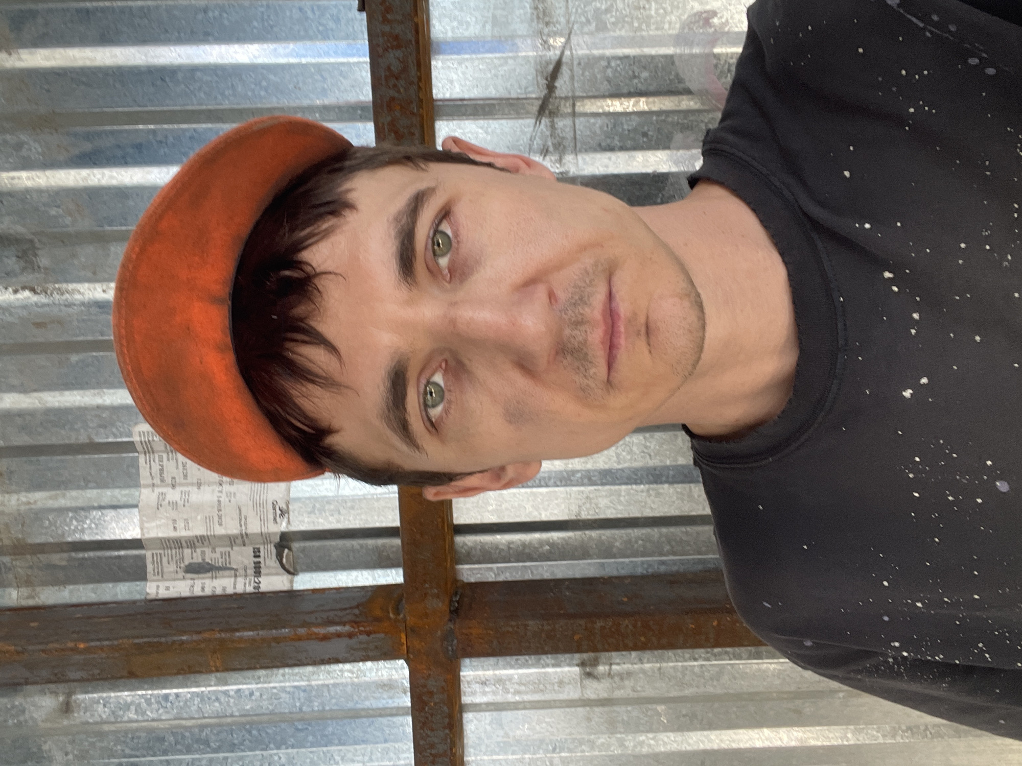 A man wearing an orange beret and a black shirt splattered with white paint is shown in a close-up selfie. He has green eyes and dark hair. His face has smudges of dirt or paint. He is standing in front of a corrugated metal wall with vertical ridges. There is a rusted metal structure on the left side of the frame, partially obscuring the wall. A crumpled piece of paper with text is attached to the metal wall behind him. The overall environment appears to be an outdoor or industrial setting. The location is identified as the city of Сокуры, Russia.