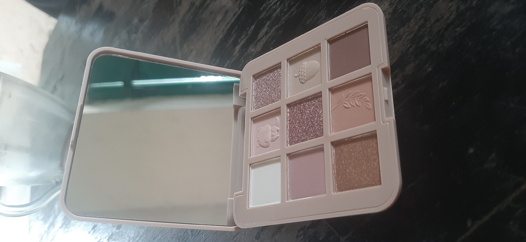 An open, light pink eyeshadow palette with nine pans is displayed on a dark, textured surface. The palette has a large mirror on the left lid. The eyeshadow pans contain a mix of matte and shimmery shades, with some pans featuring embossed designs of a squirrel, an acorn, and a leafy branch. The palette is angled, with the top left corner pointing towards the viewer. The background is blurred, showing a glimpse of a clear glass object with a white substance inside. The lighting is diffused, casting soft shadows. The scene is indoors, likely in a residential setting. The location is Yessentukskaya, Russia.