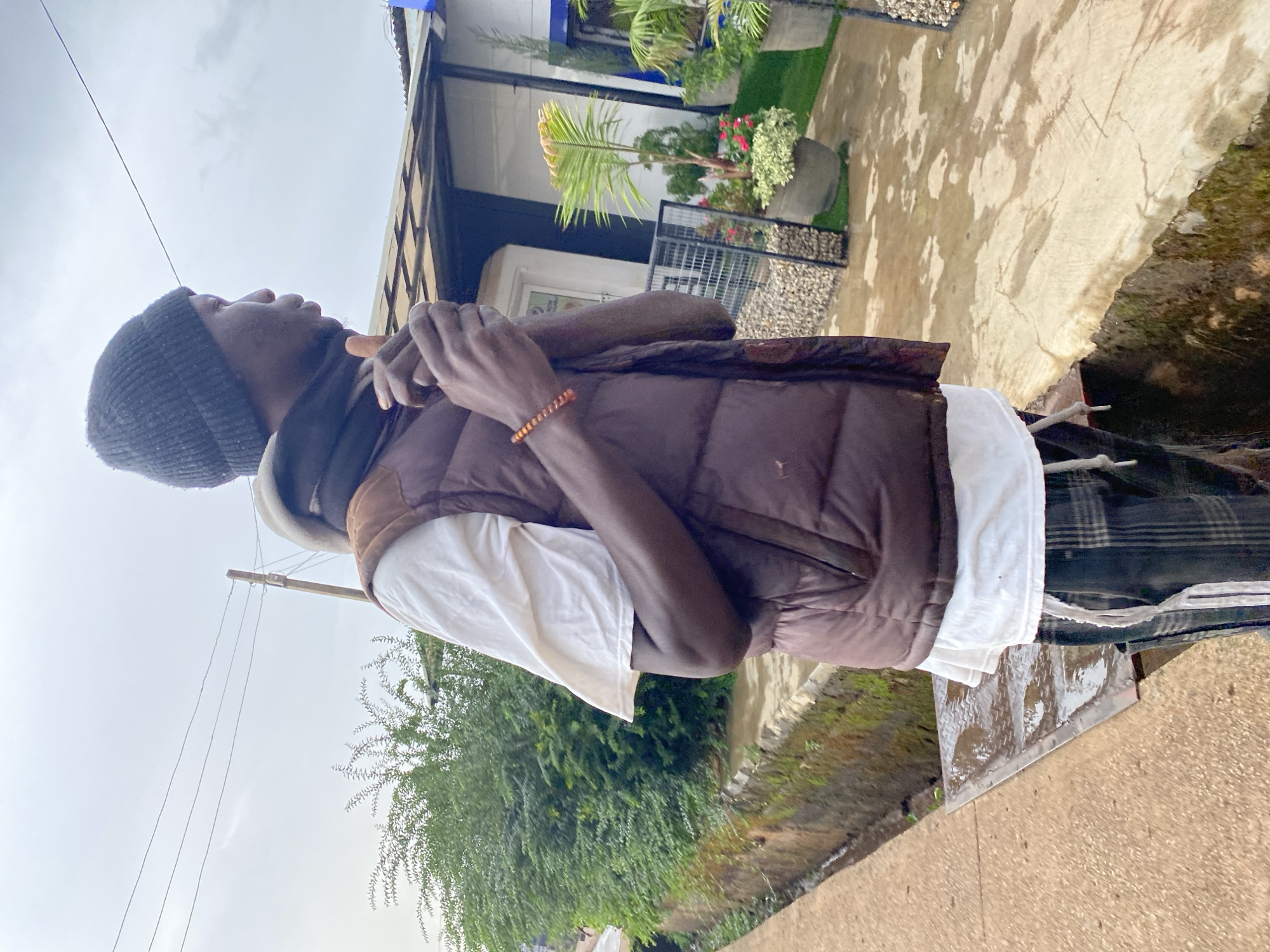 A person stands outdoors near a street, clad in a black beanie, a white t-shirt, and a brown vest. A bracelet is visible on one of their wrists. The location is Bukuru, Nigeria, as indicated by visual cues. Behind the individual is a building with plants and greenery. The environment includes a street with vegetation and a visible sky. Overhead power lines are also visible.