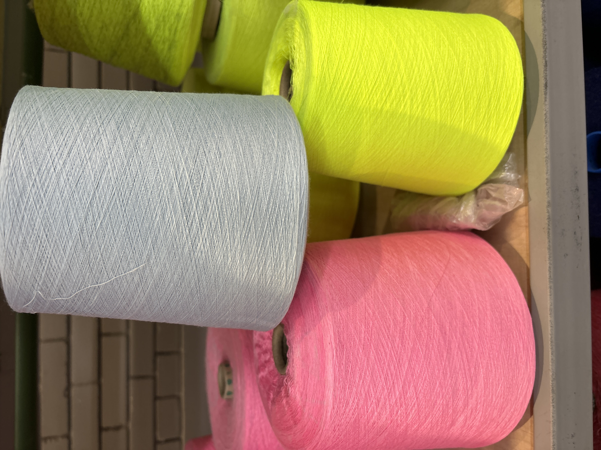 The image displays spools of thread in a storage area. The spools are stacked on shelves and vary in color, including light blue, pink, and neon yellow. The background shows a brick wall, suggesting an industrial or workshop setting. There are no people or animals visible in the image. The lighting is overhead, casting some shadows and highlighting the textures of the thread. The image does not provide any information about its location in Chaikovsky, Russia.
