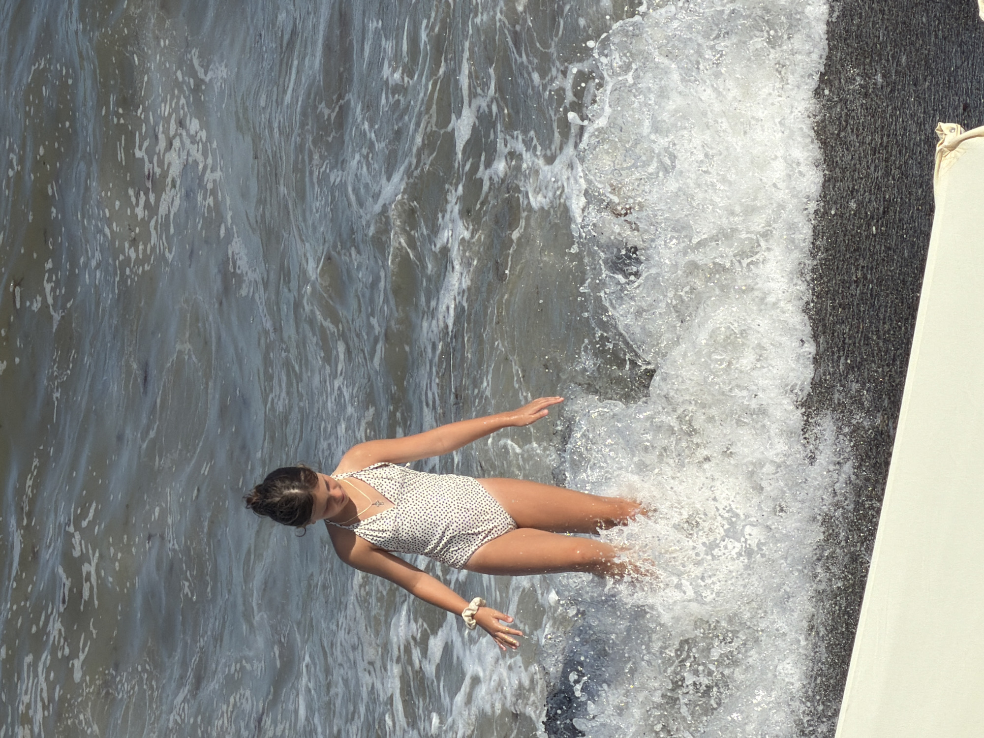 This media file shows a young woman in a speckled white swimsuit sitting in the ocean near a dark, textured surface, possibly a pier or seawall. She is positioned in the water with waves splashing around her legs and feet. Her hair is pulled back in a bun, and she is looking down, appearing to be playing or relaxing in the water. A light-colored object, perhaps a life preserver or part of a boat, is visible on the right side of the frame. The water is a mix of dark and light tones, with white foam from the waves. The scene suggests a sunny day, as indicated by the bright light and reflections on the water. The location is identified as Alushta, Ukraine. No visible text is present in the media file.