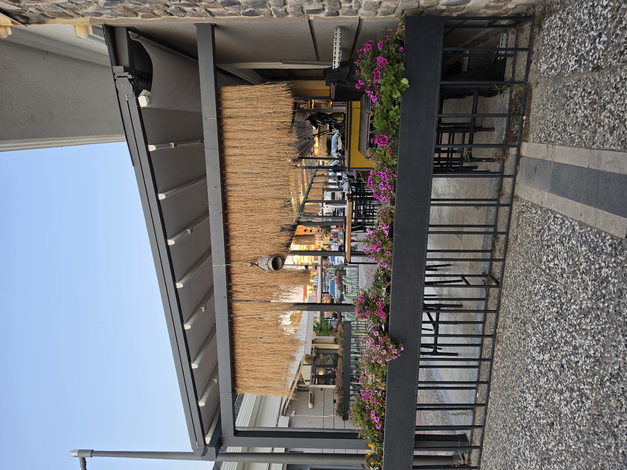 The image shows an outdoor seating area of a restaurant or cafe. The seating area is covered by a large awning with a thatched roof, supported by dark metal beams. In the foreground, there is a black metal planter box filled with vibrant pink and green flowers, adding a splash of color to the scene. Behind the planter box, rows of tables and chairs are visible, suggesting that the establishment is open for business. Some people can be seen in the background, either seated at tables or walking, indicating a daytime setting. The ground is paved with small, irregularly shaped stones, a common feature in Mediterranean settings. The overall atmosphere appears relaxed and inviting, with the bright flowers and the open-air setup suggesting a pleasant day. The building behind the awning has a stone facade, and the sky is clear and blue, indicating fair weather. There is no visible text in the image.