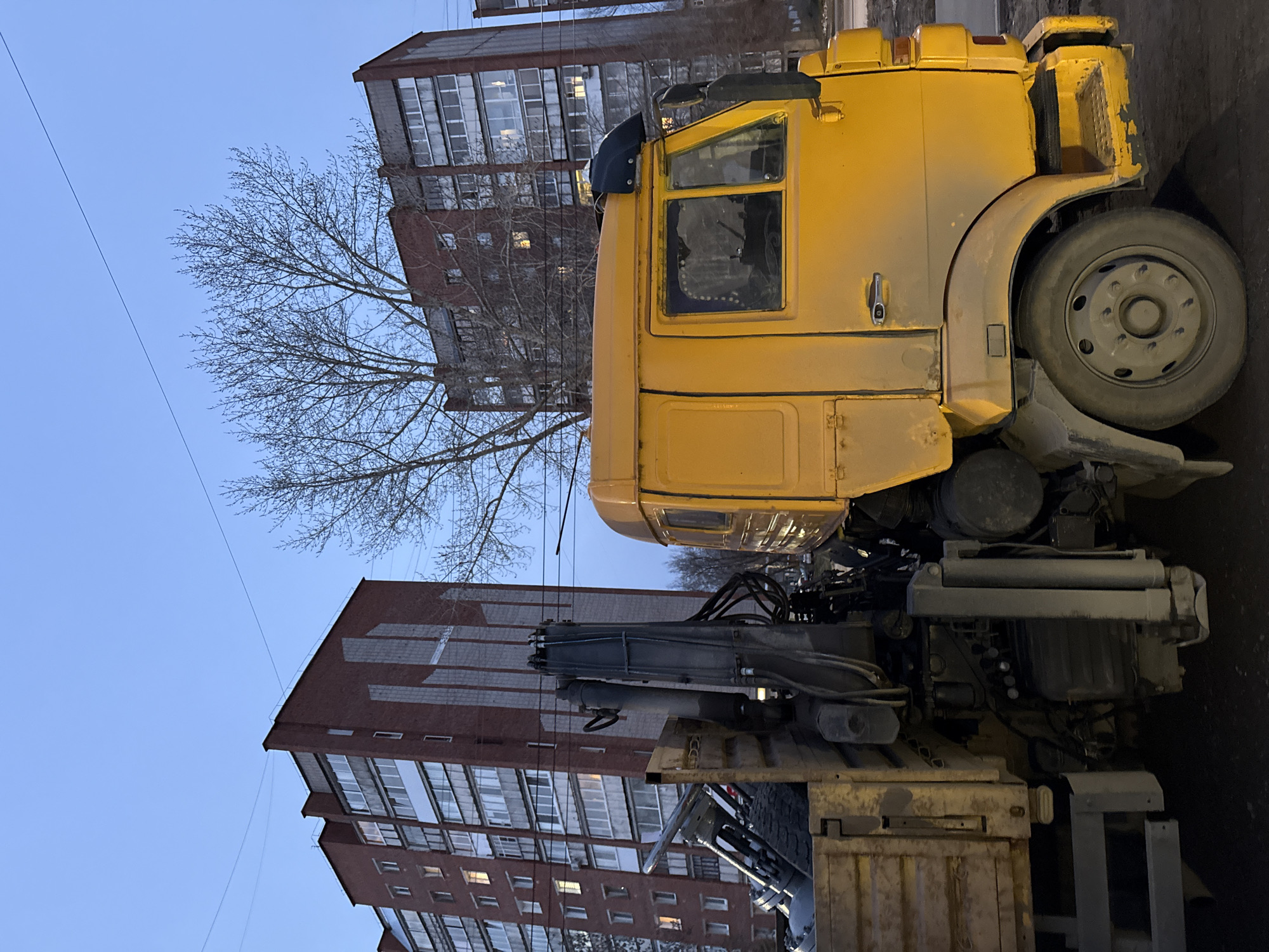 A yellow KamAZ truck is parked in Votkinsk, Russia. The truck is positioned on a city street next to a building made of red brick. The sky is light blue. A tree, bereft of leaves, is visible in the background. The crane mechanism is mounted on the back of the truck.