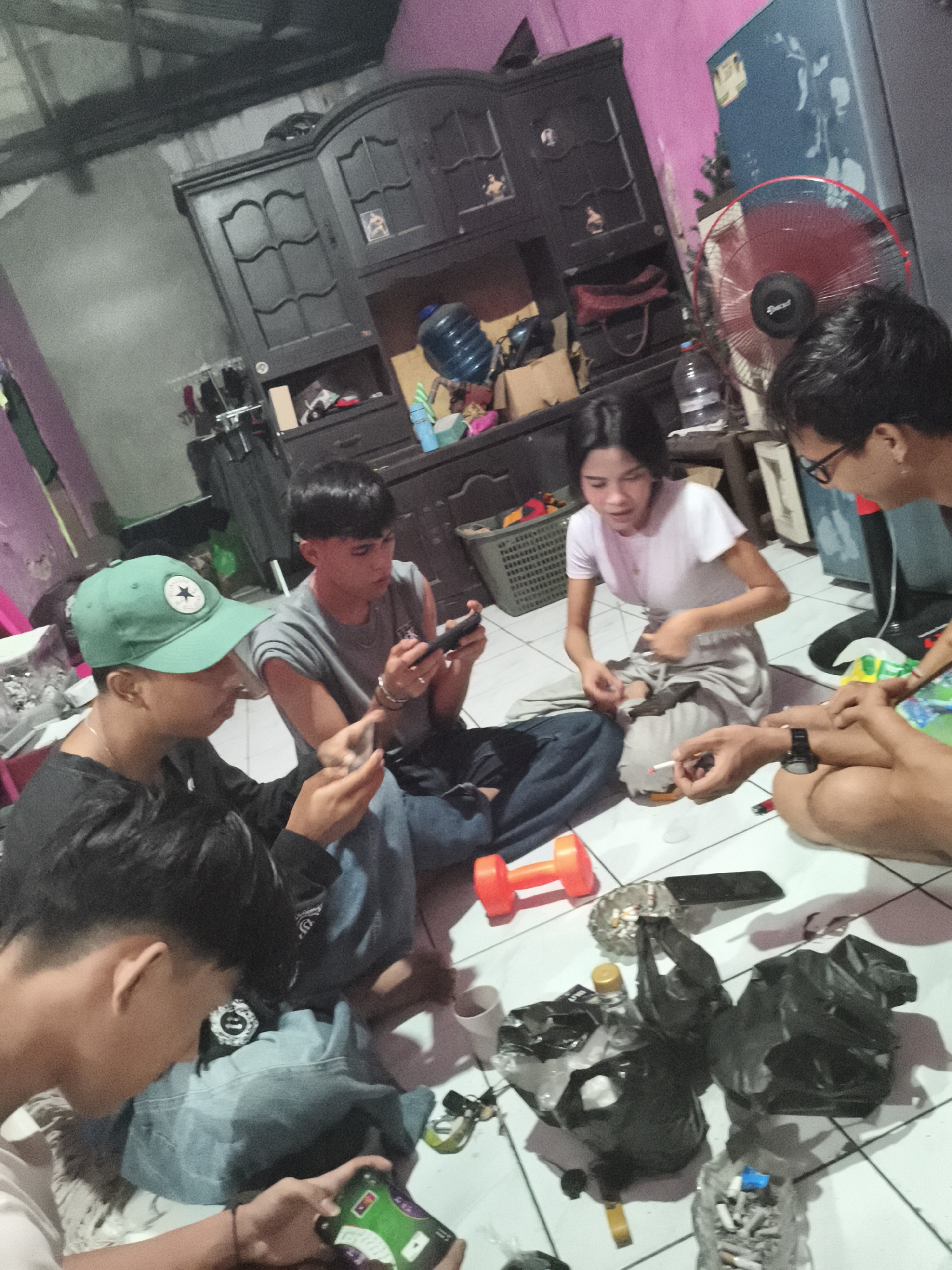 The image depicts a group of young people, likely teenagers or young adults, gathered indoors on a tiled floor, possibly in Depok, Indonesia. The scene appears informal, with individuals sitting or kneeling around a central area. Several people are actively engaged with their mobile phones, suggesting a shared activity like gaming or social media browsing.
In the foreground, a person wearing a green baseball cap is looking down at a phone in their hands. To their right, another individual in a grey sleeveless shirt is also focused on a phone. Further to the right, a young woman in a white t-shirt is seated, with what appears to be a cigarette in her hand and a small white object on the floor beside her. Another person is visible to the far right, wearing glasses and looking towards the center of the group.
The environment includes a dark wooden cabinet with glass doors in the background, a large water jug, and a red oscillating fan. Several black plastic bags, some containing what look like trash, are scattered on the floor. Other objects visible include an orange dumbbell, an ashtray filled with cigarette butts, and various small items. The lighting appears to be artificial, contributing to a somewhat dim and intimate atmosphere. The overall impression is one of a casual social gathering, possibly in a residential setting.