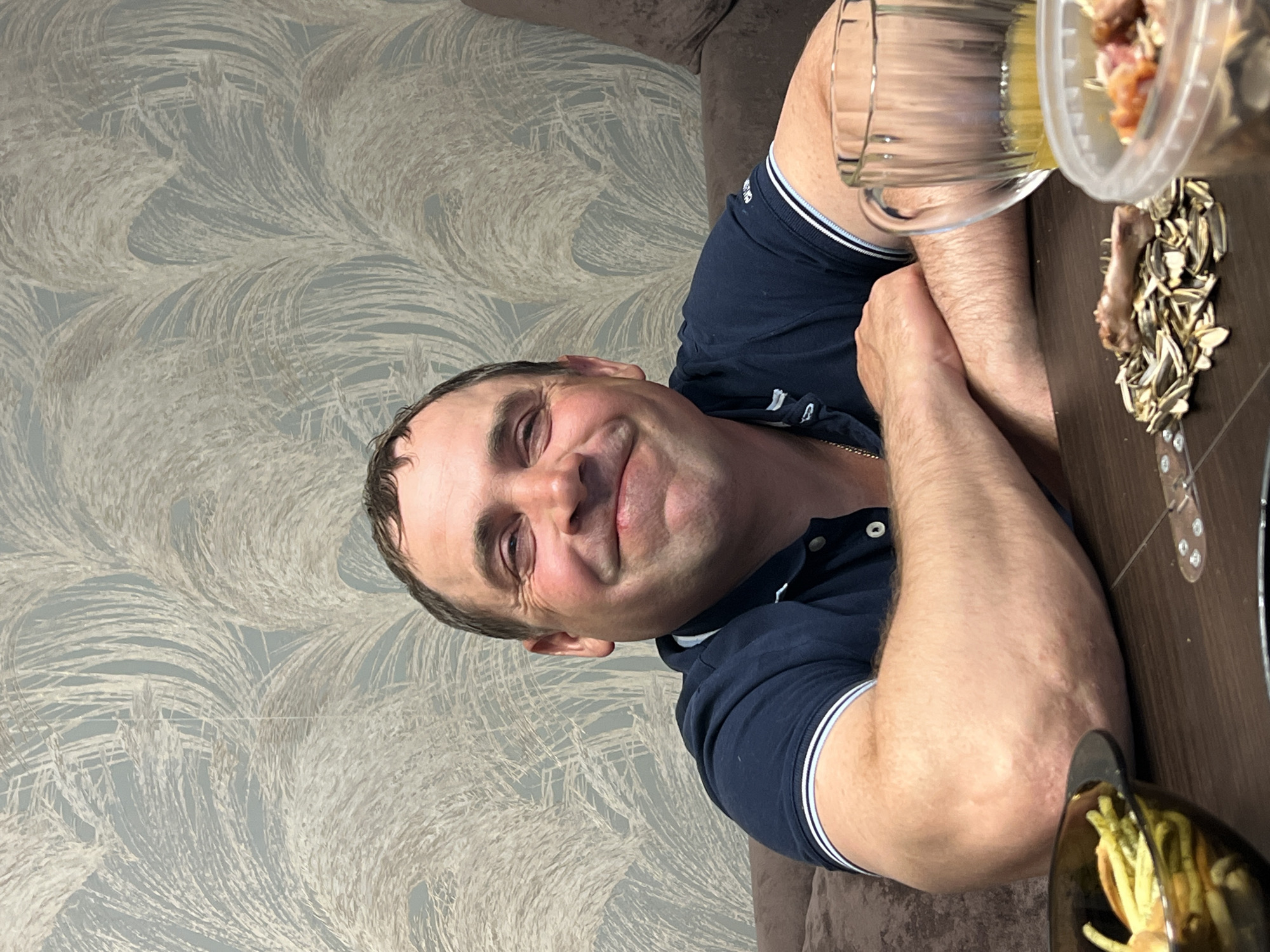 A person is photographed indoors at a table with food and drink. The person is a middle-aged man with short dark hair and fair skin, wearing a dark blue polo shirt with white trim at the collar and sleeves. He is smiling at the camera. On the table, there are multiple containers with food, including sunflower seeds and what appears to be salad. There's a glass mug or container possibly containing a yellowish liquid. The wall behind him is covered in wallpaper with a textured, wave-like pattern. The location is identified as Noviy, Russia.