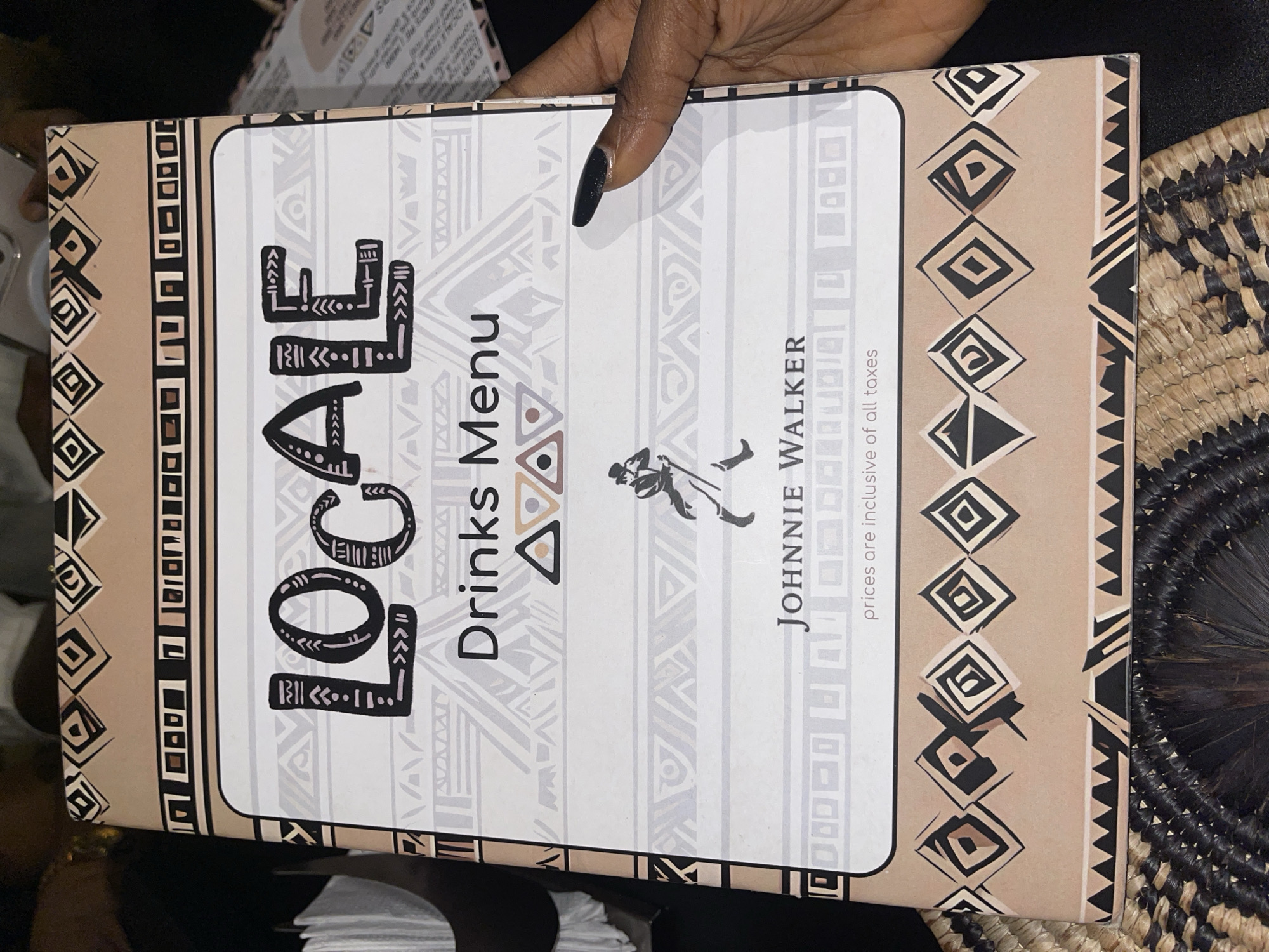 A cropped indoor shot presents a "Drinks Menu" cover, emblazoned with "LOCALE" in bold, stylized lettering. The menu is a paper booklet with decorative borders of ethnic-inspired patterns, held by a person with manicured nails. A "JOHNNIE WALKER" logo is visible, and the phrase "prices are inclusive of all taxes" is printed on the cover. The menu indicates that "LOCALE" is located at Kamata Rode in Ikoyi, Nigeria. A woven placemat appears in the background on the right side of the image.