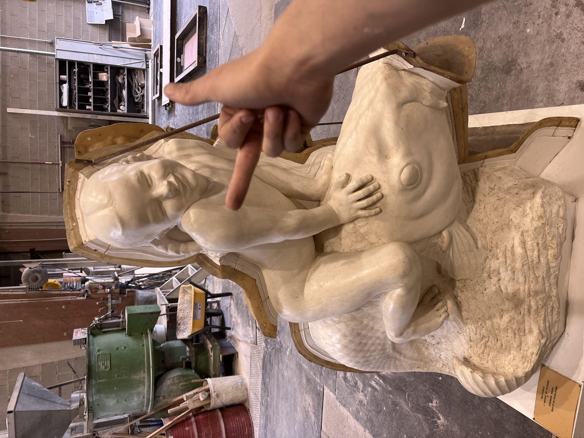A partially-visible hand points with its index finger towards a white sculpture resting in a work area. The sculpture depicts a human figure lying atop a fish, both embedded within what appears to be a plaster mold. The sculpture and mold are predominantly off-white in color. The surrounding environment is cluttered with equipment and materials.
Located in Saint-Rémy-lès-Chevreuse, France.