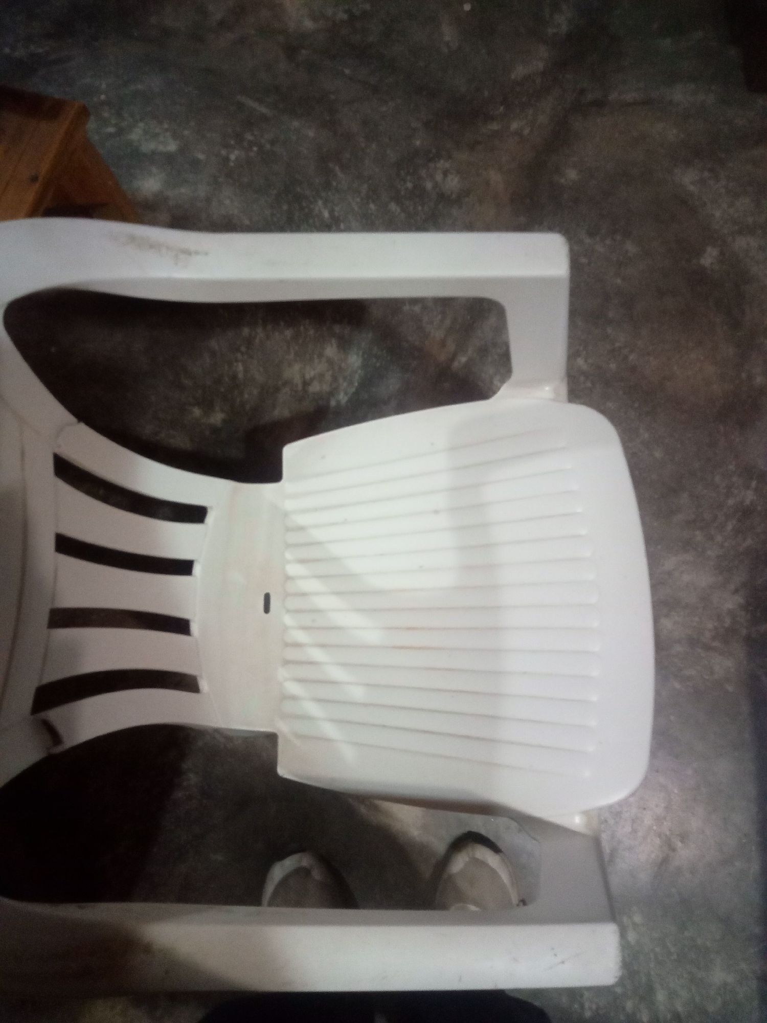 The image shows a white plastic chair from a top-down perspective, placed on a textured, dark gray concrete floor. The chair has a slatted backrest and a textured seat. A small portion of a wooden object, possibly a table or cabinet, is visible in the upper left corner of the frame. Two feet wearing light-colored shoes are partially visible underneath the chair, indicating someone is sitting on it. The overall lighting suggests an indoor environment. The location is identified as Altagracia, Venezuela.