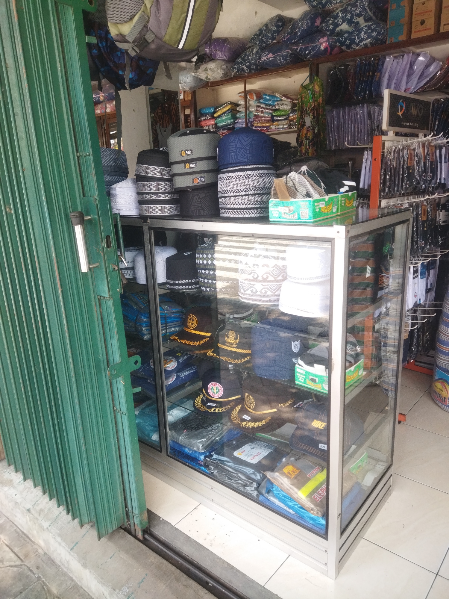The image shows a small retail stall in the city of Wonogiri, Indonesia, specializing in Islamic caps, known as peci or songkok. The stall features a glass display cabinet filled with various styles and colors of these caps, neatly stacked on shelves. Some caps have intricate patterns, while others are plain. In addition to caps, the cabinet also contains some folded fabrics, possibly sarongs or other religious attire, and a few packaged items.
To the left of the cabinet, a large, green, corrugated metal accordion door is partially closed, obscuring some of the stall's interior. The door has a metal handle and appears to be the main entrance or storefront. Above the door, partially visible, are stacks of bags and textiles, suggesting further merchandise.
The background reveals the interior of the shop, with shelves stocked with more clothing items, including what appear to be shirts and other garments, neatly hung or folded. There's a sign with text that is difficult to read but seems to indicate branding or information about the products. The flooring is tiled with light-colored square tiles. The overall impression is of a modest, local shop selling religious and casual wear.