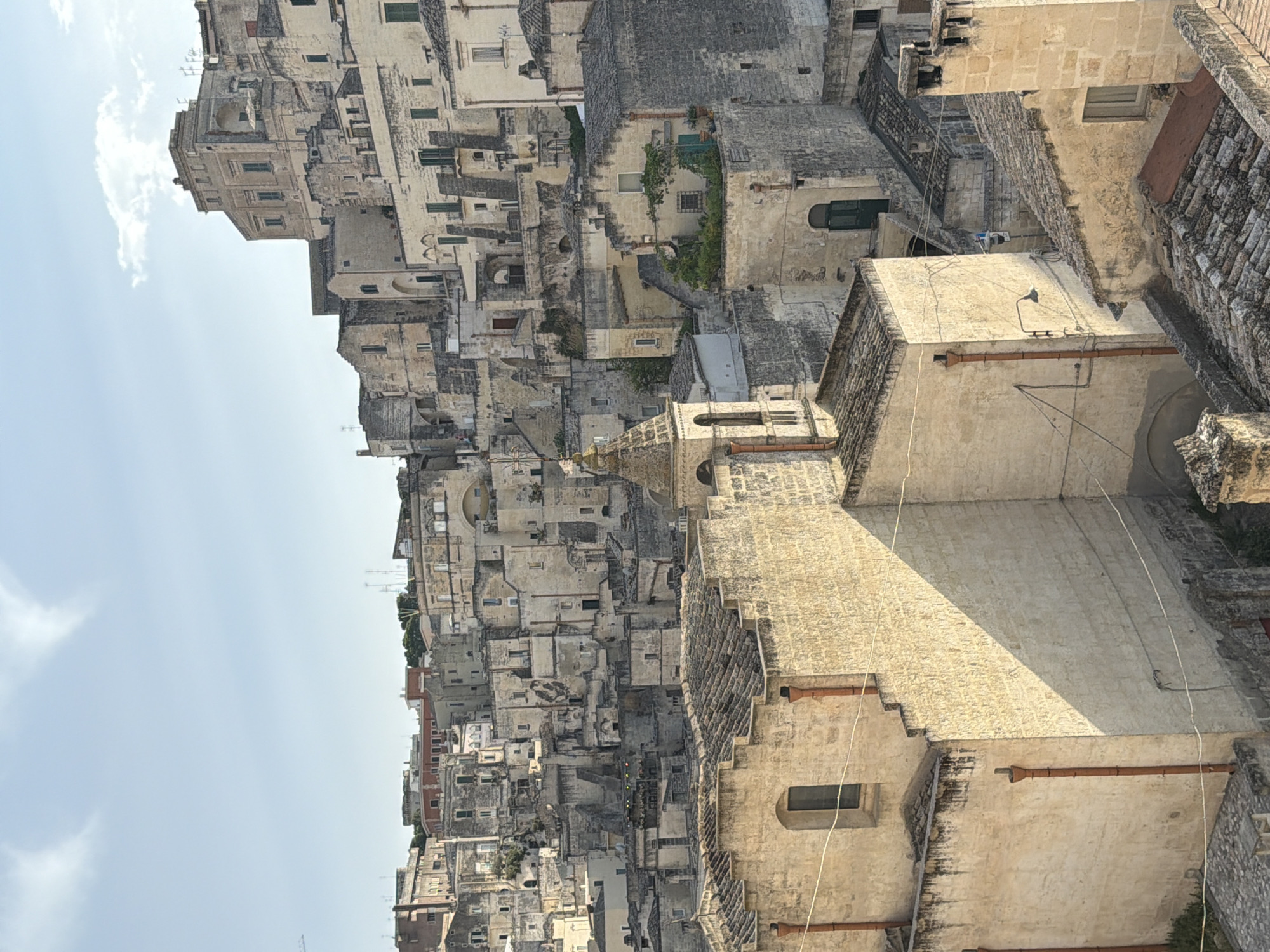 The image shows a dense urban landscape of ancient stone buildings in Matera, Italy. The architecture is characterized by tightly packed structures, many with cave-like dwellings carved into the rock, and traditional tiled roofs. The color palette is dominated by earthy tones of beige and gray, reflecting the local Sassi di Matera. The sun casts long shadows, suggesting it is either early morning or late afternoon. There are no visible people or animals. The scene is one of historical architecture and urban density.