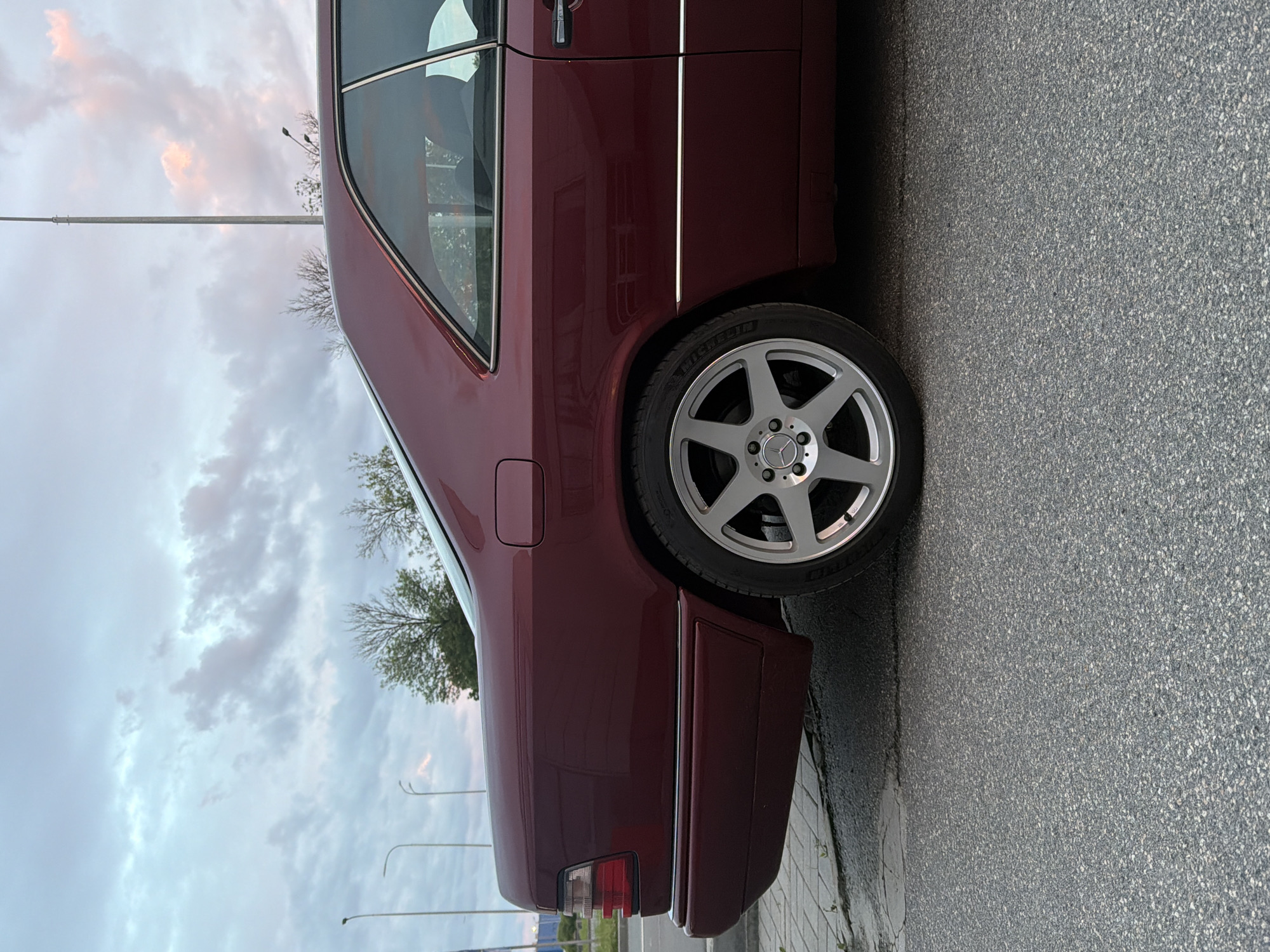 This is a medium shot of the rear passenger side of a maroon Mercedes-Benz coupe parked on an asphalt road. The car has a chrome trim line running along the lower body. The car's rear wheel, a silver alloy with five spokes and a Mercedes-Benz logo in the center, is visible. The tire is a Michelin. The background shows a cloudy sky with some patches of pink and orange from the sunset. There are streetlights and bare trees visible in the distance, along with a sidewalk made of interlocking pavers. The overall scene appears to be in a suburban or urban setting.