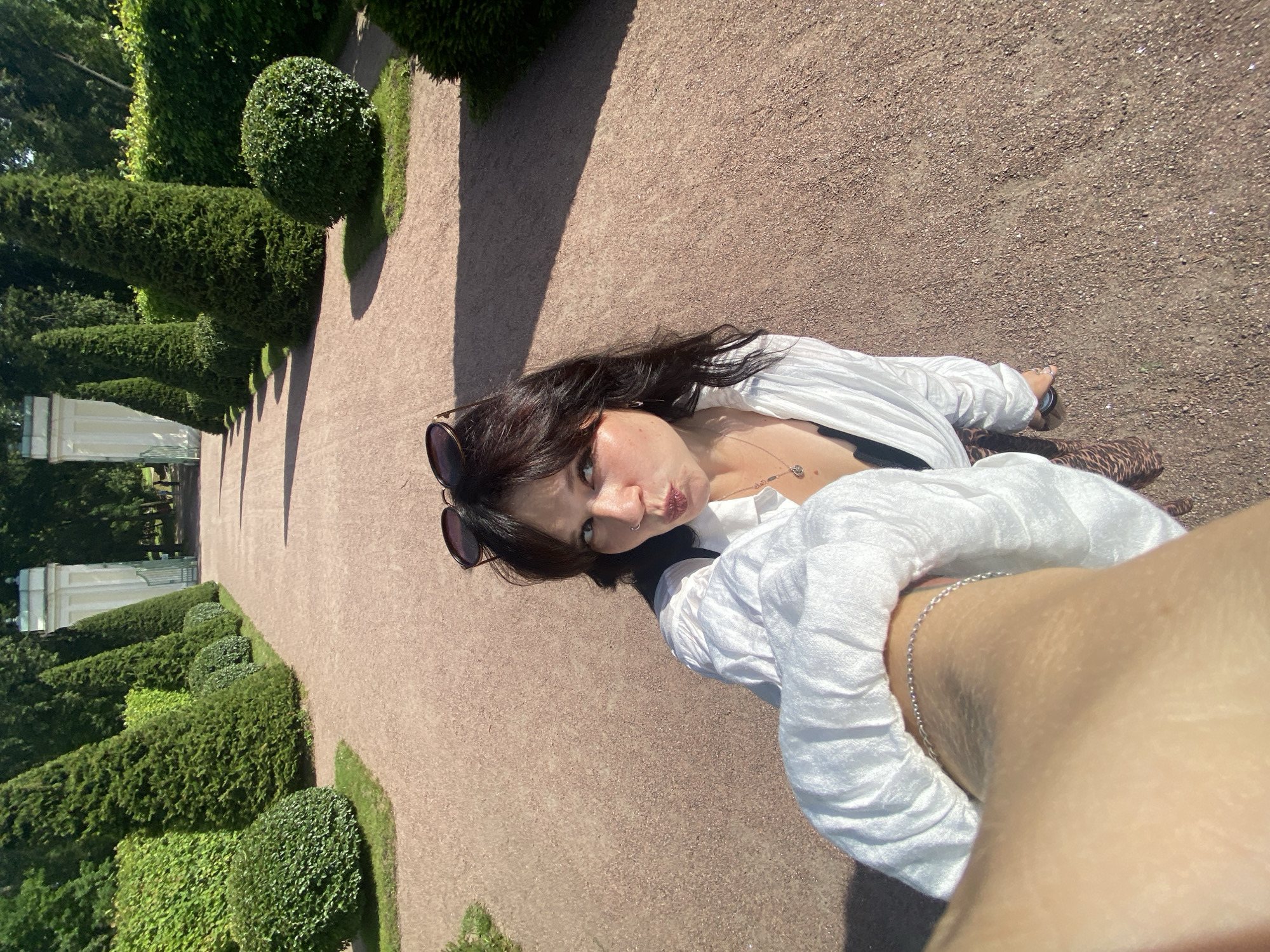 An individual is taking a selfie while walking on a gravel path in the park of the Oranienbaum ensemble in Lomonosov, Russia. The individual is wearing a white, open-collared shirt, a black top underneath, and patterned leggings. Sunglasses are perched on their head. The path is lined with manicured topiaries and dense green hedges. In the background, white classical pavilions frame a view down the path, with tall trees forming a dense backdrop. The sunlight casts distinct shadows across the path and the surrounding greenery. The scene is set in the summer, indicated by the lush vegetation and clear daylight.
