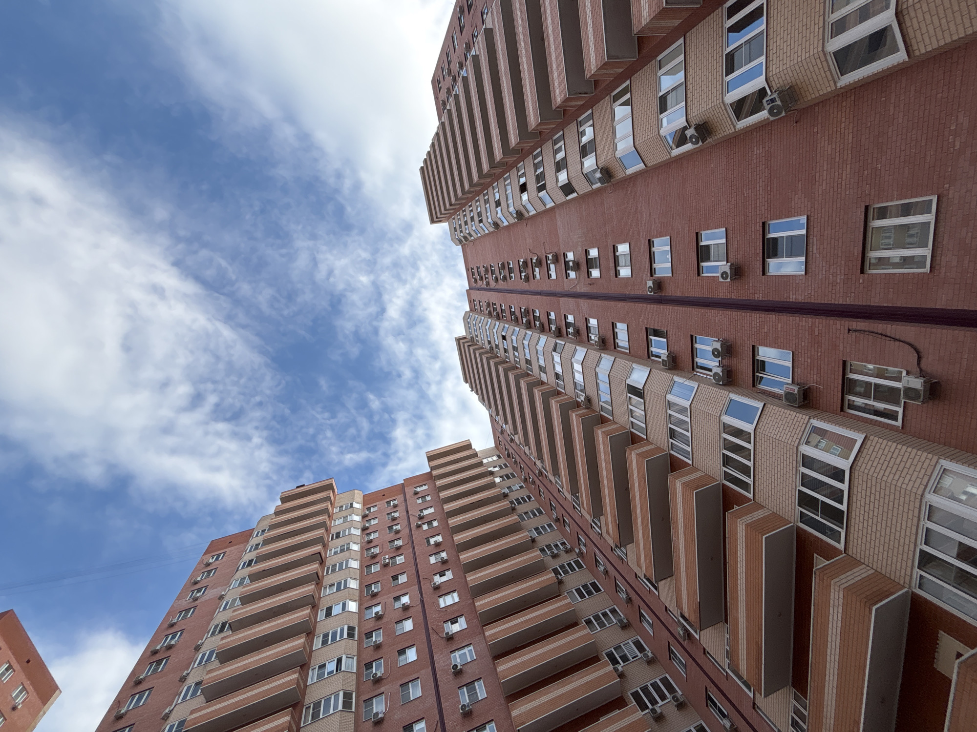 This media file is an upward-looking shot of two tall, multi-story apartment buildings constructed with red brick. Both buildings feature numerous balconies with reddish-brown railings, and many windows with white frames. Air conditioning units are visible mounted on the exterior walls below many of the windows. The sky above is blue with scattered white clouds, suggesting it is daytime. The perspective makes the buildings appear to lean into the sky. The buildings are modern in appearance and appear to be residential. No people are visible. The scene is likely a residential area in Krasnodar, Russia.