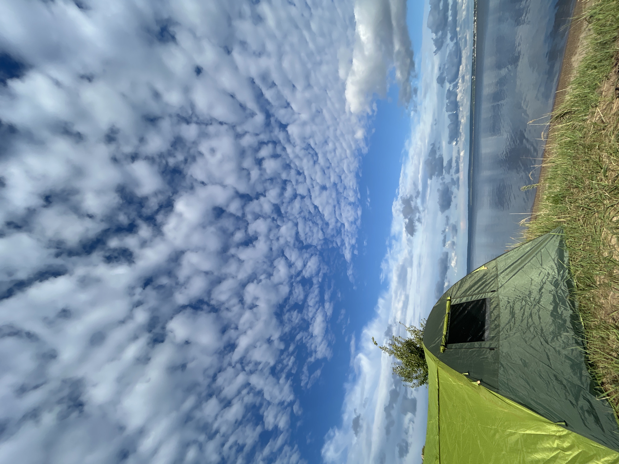 The image shows a green tent pitched on a grassy bank overlooking a wide body of water, likely a river or lake, in Noviy, Russia. The sky is filled with a dramatic display of fluffy white and gray clouds against a bright blue background. The water reflects the sky and the distant shoreline, where a town or settlement is visible. The tent is partially in the foreground, with its entrance and a small window visible. The grass is tall and green, suggesting a natural, possibly wild setting. The overall scene conveys a sense of peaceful outdoor recreation.