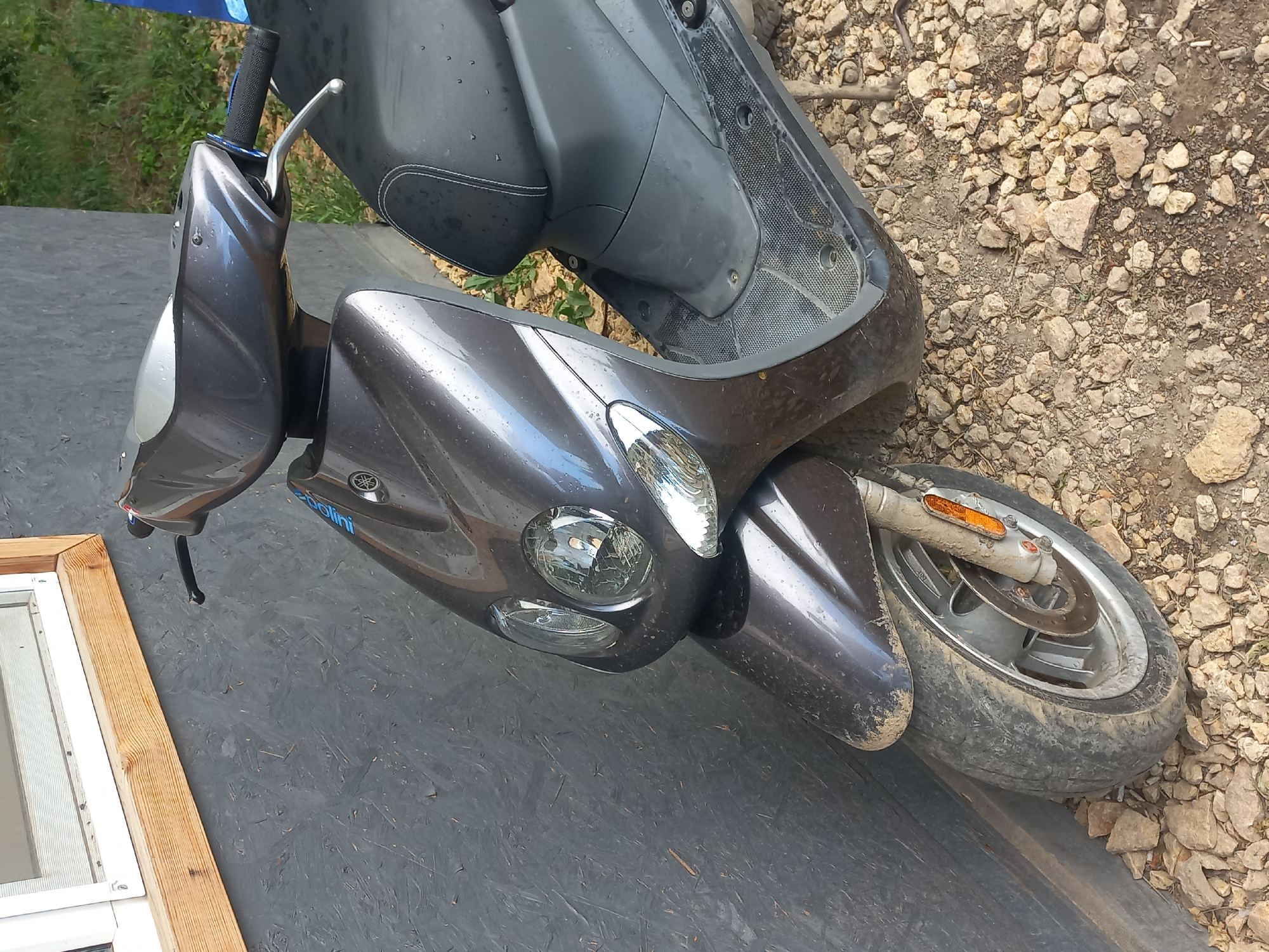 A dark metallic gray scooter, bearing a Yamaha logo and a blue "Polini" decal on its front fairing, is parked in the city of Velykodolynske, Ukraine. The scooter's body panels display numerous water droplets, while its front fender, tire, and wheel are coated with light brown dust and dirt. Visible components include dual circular headlights, clear front turn signal lenses, a black seat with white stitching, and a textured black plastic floorboard. The left handlebar is equipped with a black grip featuring blue accents and a silver brake lever. The front wheel assembly incorporates a disc brake and an orange reflector on the fork. The vehicle is positioned adjacent to a dark gray wall with a coarse, layered texture. A white-framed window with a natural wood sill is partially visible above the wall. The ground beneath the scooter consists of scattered light-colored gravel and packed earth. Green foliage is visible in the background to the right, partially obscuring a blue object.