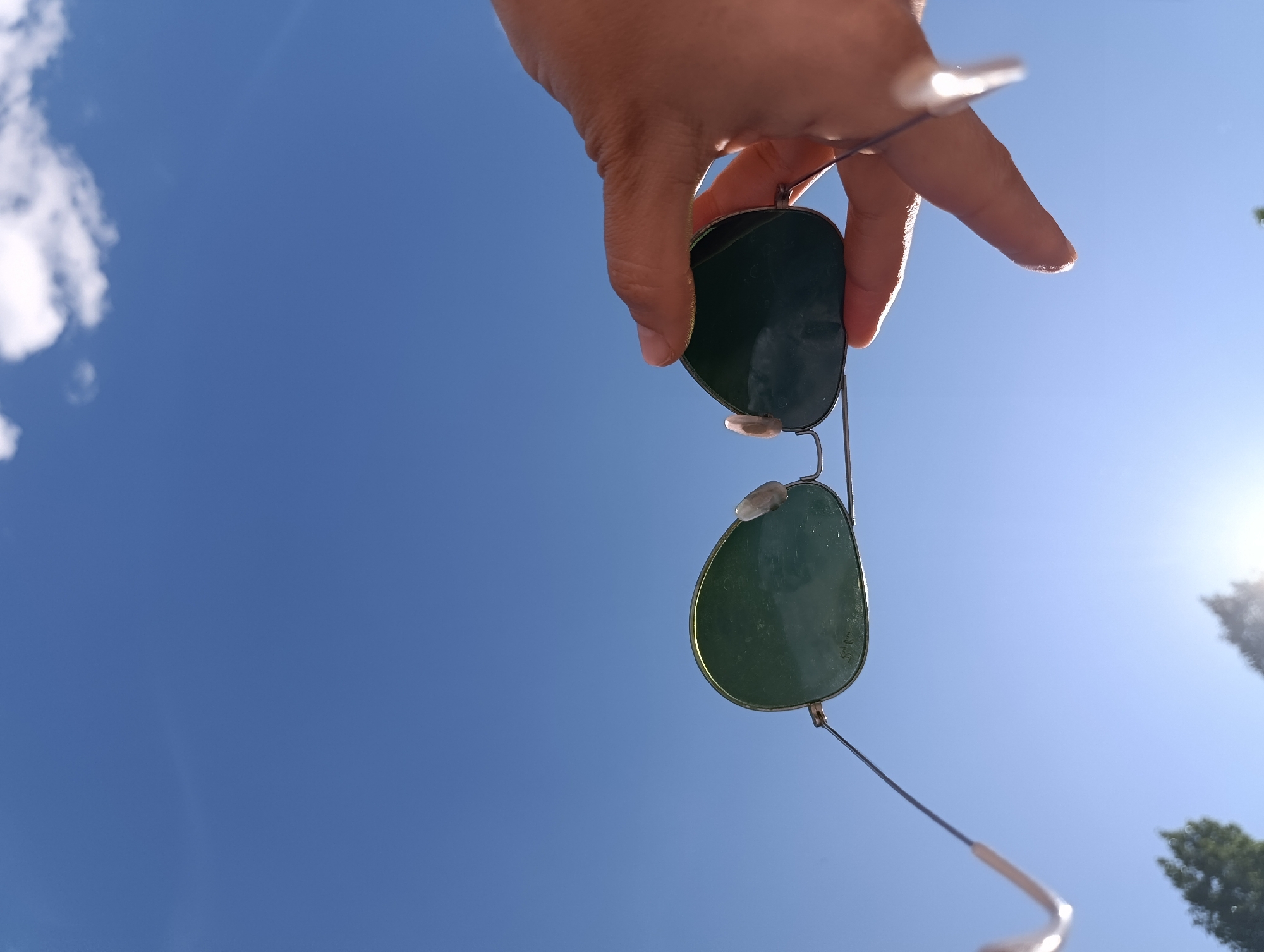 A hand holds a pair of aviator sunglasses, with their green lenses facing downwards, against a clear blue sky. The sunglasses are suspended by a thin string or wire, held between the thumb and forefinger of the hand. The hand exhibits long, manicured fingernails, painted with a light color. The background consists of a vast expanse of blue sky, with a few wispy white clouds visible on the left side and a bright sun shining in the upper right corner, causing some lens flare. The perspective is from below, looking up towards the sky. The scene does not contain any identifiable landmarks or elements that would place it specifically in Saratov, Russia; it is a generic outdoor scene.