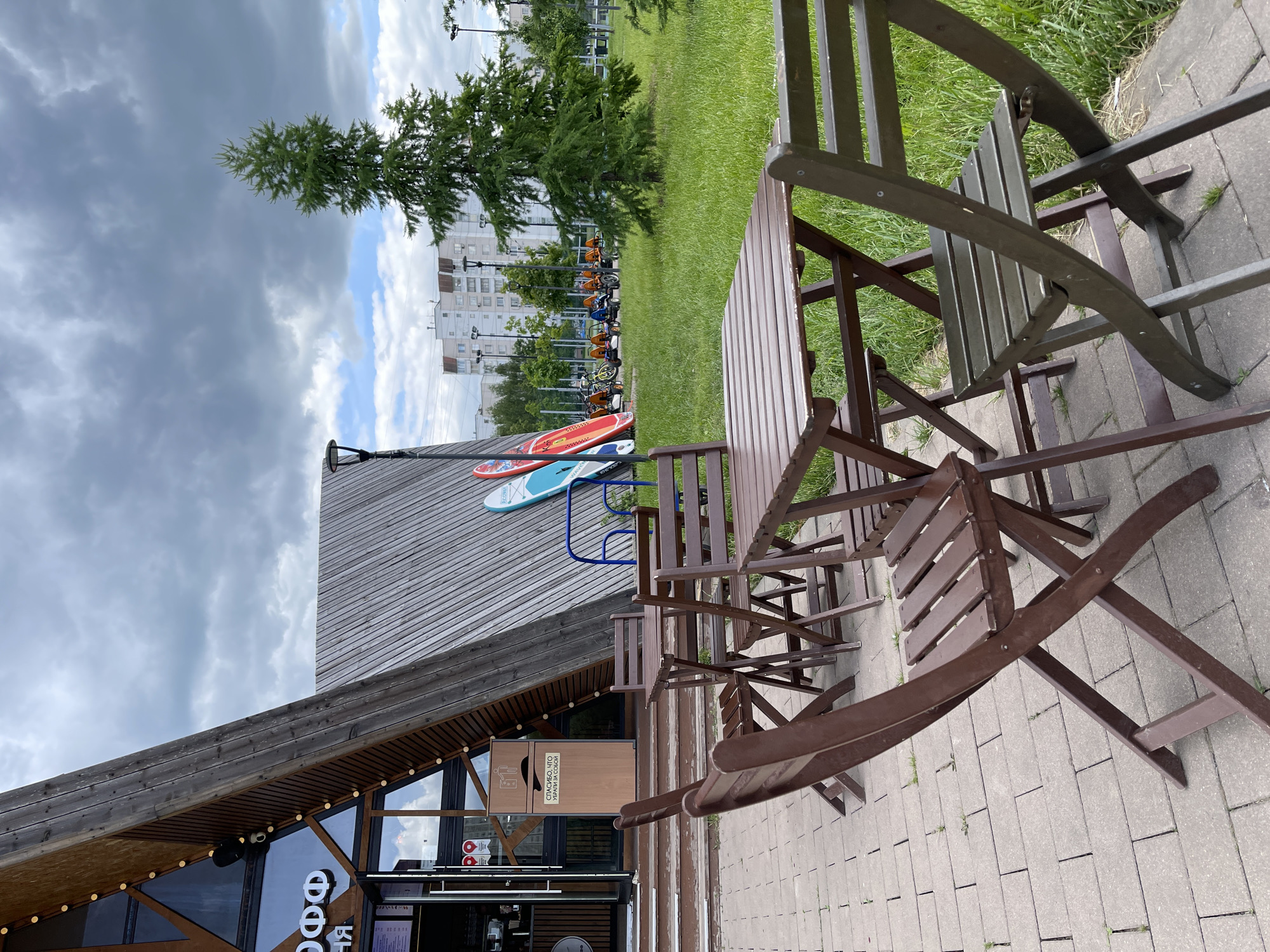 This image depicts an outdoor scene in Moscow, Russia. A wooden building with an A-frame roof and a glass front is visible. The building appears to be a cafe, as evidenced by the signage. Outside the cafe, there are wooden tables and folding chairs arranged on a paved area. On the roof of the building, there are several paddleboards and a few tricycles. In the background, there is a grassy area, a line of tricycles parked, a few trees, and an apartment building. The sky is cloudy. There are no people visible in the image.