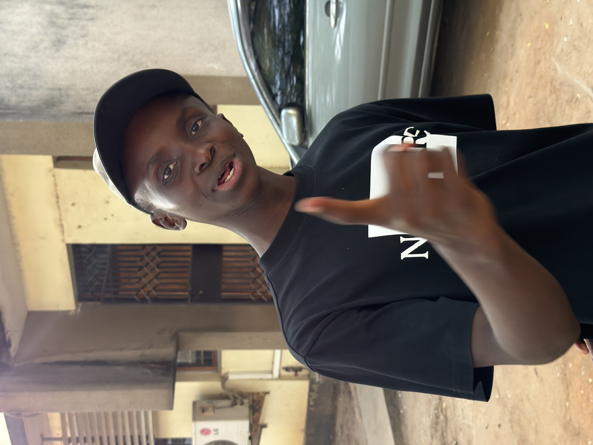 In Dar es Salaam, Tanzania, a young Black male, wearing a black t-shirt with white lettering and a black baseball cap, stands outdoors. He is looking towards the camera with his mouth slightly open and his right thumb raised in a "thumbs-up" gesture. Behind him, a light-colored car is parked, and part of a building with a yellow facade, a window with bars, and an air conditioning unit is visible. The ground is sandy.