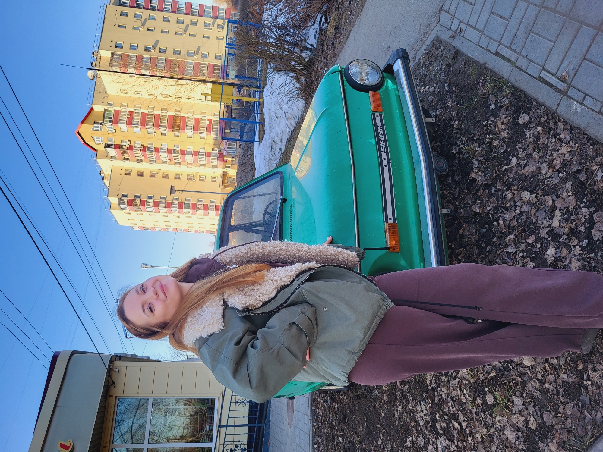 A woman stands in front of a green ZAZ-968M car in Chaikovsky, Russia. She is wearing a green jacket with a light-colored fuzzy lining, a dark top, and maroon sweatpants. The car is parked near a sidewalk with fallen leaves. Behind the car, there is a building, possibly an apartment complex, with red and yellow details, and a blue metal fence. The sky is clear blue. Other buildings and power lines are visible in the background.