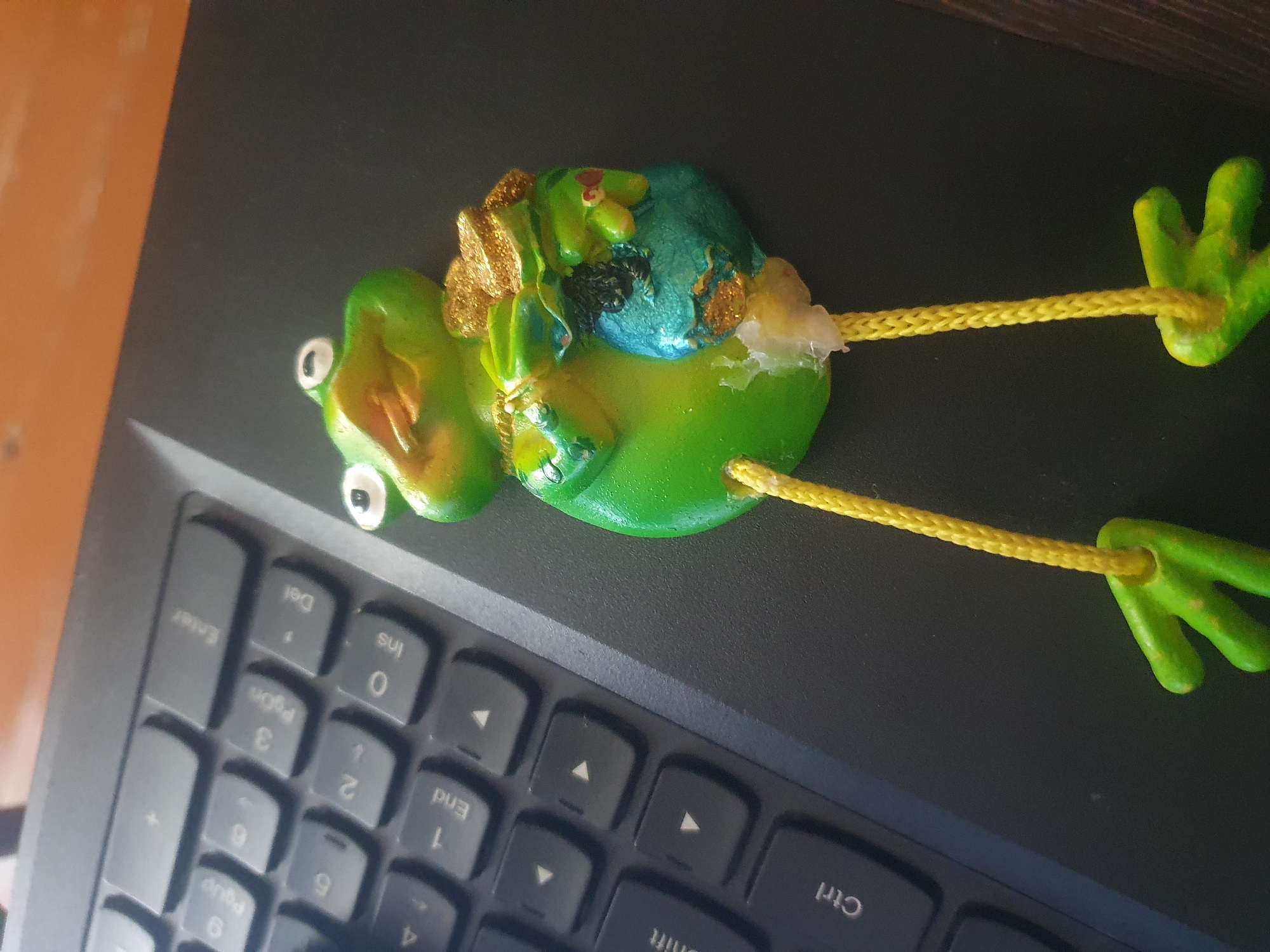 A green, frog-shaped figurine with gold accents and yellow strings is placed on a black laptop keyboard. The figurine appears to be made of resin or plastic and has a whimsical design, with oversized eyes and outstretched limbs. It is holding a small, blue pouch filled with gold glitter. The keyboard is partially visible, with some keys showing labels like "Enter", "Shift", and "Ctrl". The background consists of a wooden surface, likely a desk, and a dark, textured surface. The image is taken from a top-down perspective, with the figurine slightly off-center. The overall scene is domestic or office-like, suggesting the figurine is a decorative item placed on a workspace.