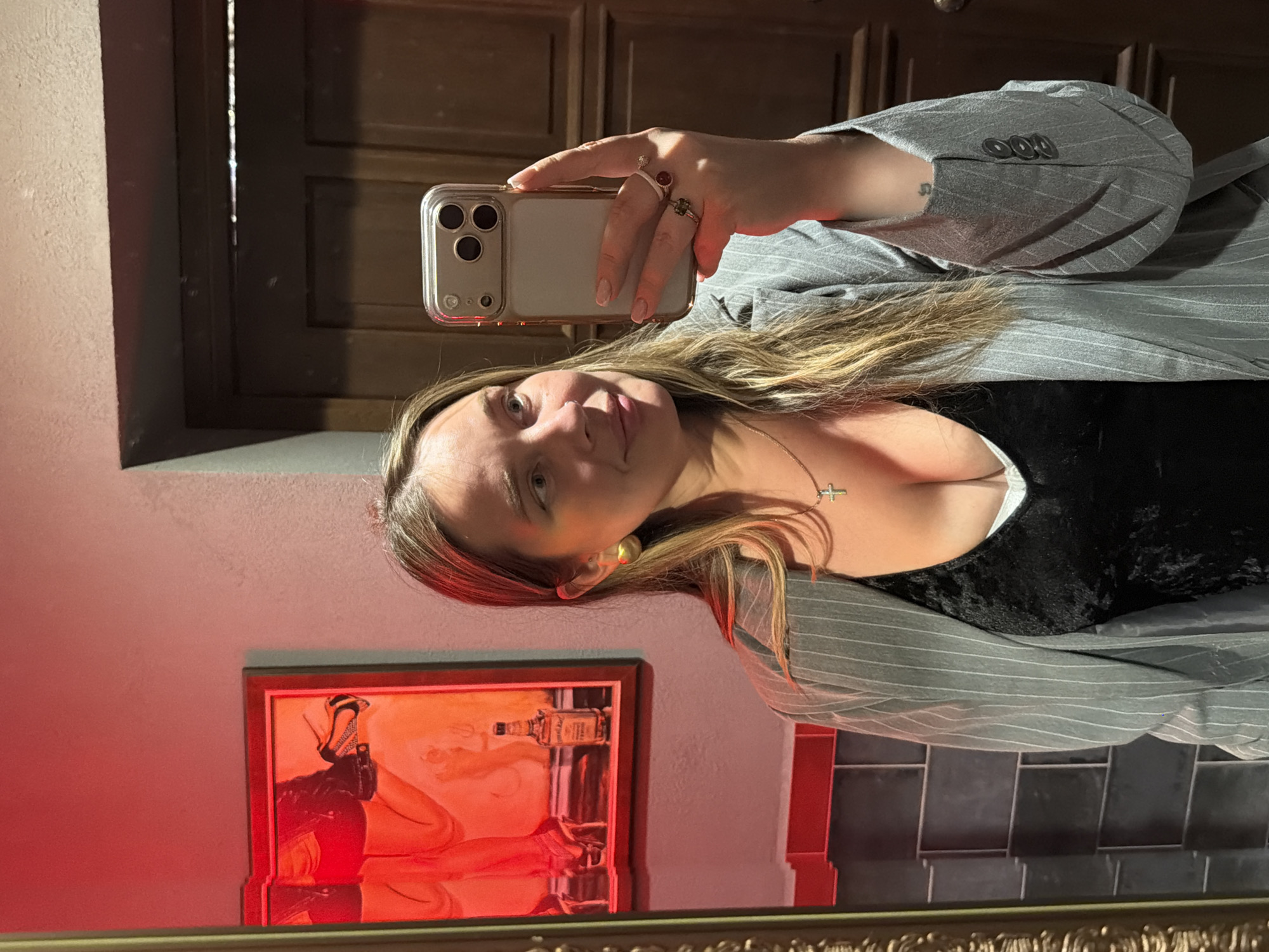 A woman is taking a selfie in what appears to be a bathroom. She is wearing a gray pinstripe blazer over a black top. A framed picture with red lighting hangs on the wall. The background includes wooden doors and dark gray square tiles. A reflection suggests the photo is taken in a mirror. There is no definitive indication of Moscow, Russia.