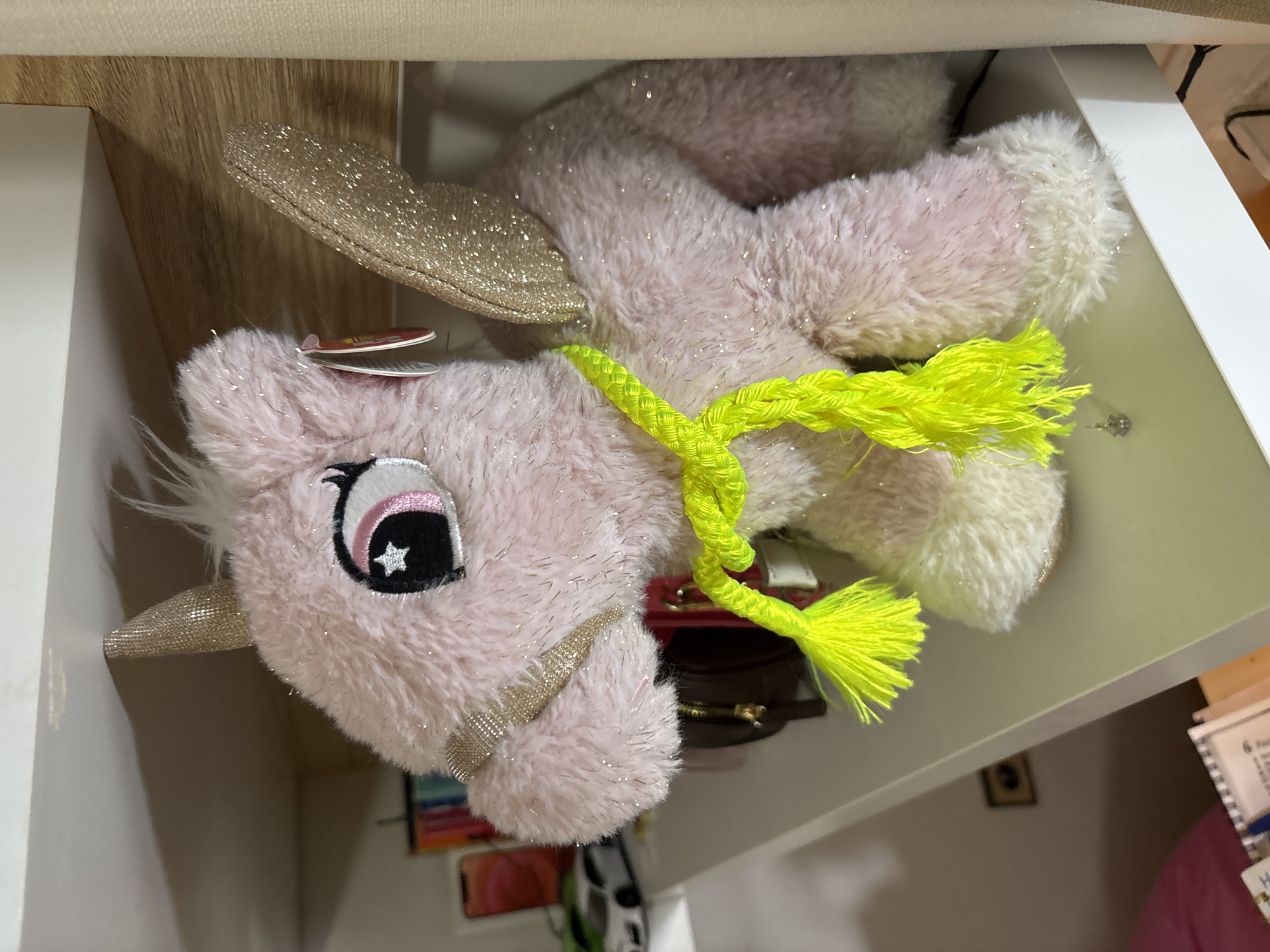 The image shows a pink stuffed unicorn with glittery wings and a gold-colored horn, inside a white storage space, possibly a drawer or shelf. The unicorn has a bright yellow braided rope around its neck. Other items are visible in the storage space, including what appears to be a phone, a wallet or small purse, and a book. The photograph's orientation is sideways. The location of the image in the city of Sofia, Bulgaria, is not provided.