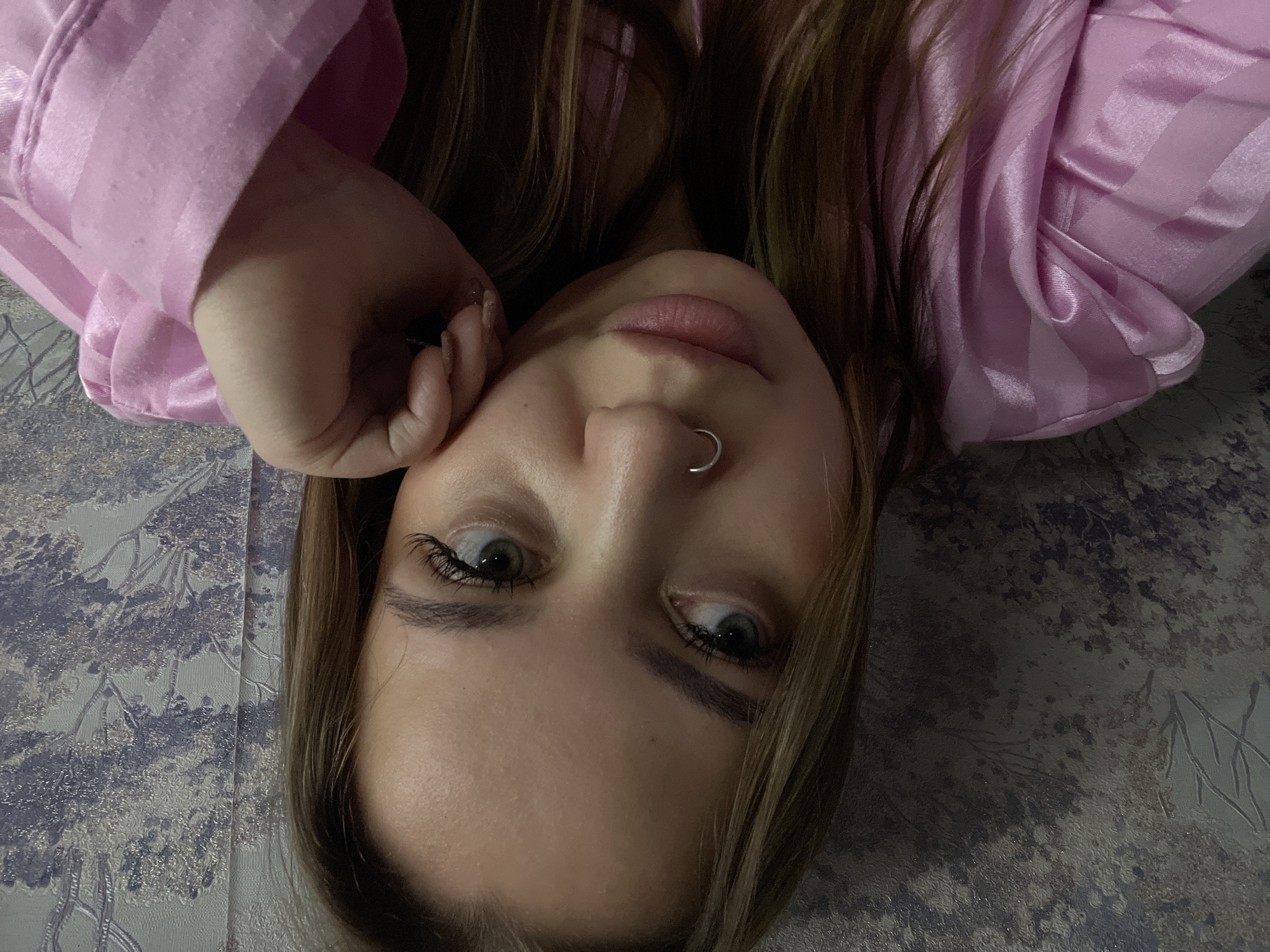 A person is lying down, facing upward at an angle. The person has light brown hair, fair skin, and light-colored eyes. A silver hoop is visible in the person's left nostril. The person is wearing a pink satin shirt or pajama top, and one hand is near the left side of the face. The background is a patterned fabric with a pale blue and gray color scheme, reminiscent of branches or trees. There is no information in the image about the location being in Novopokrovskaya, Russia.